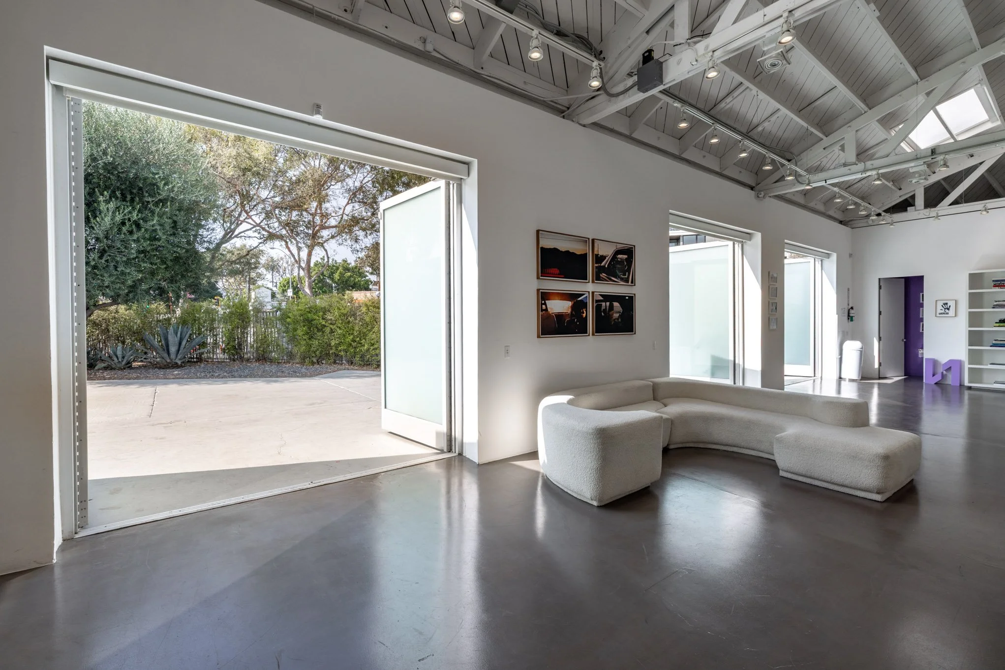 Modern art gallery with open door leading outside, white curved sofa, framed photographs on white wall, bookshelves, purple sculpture, and large windows providing natural light.