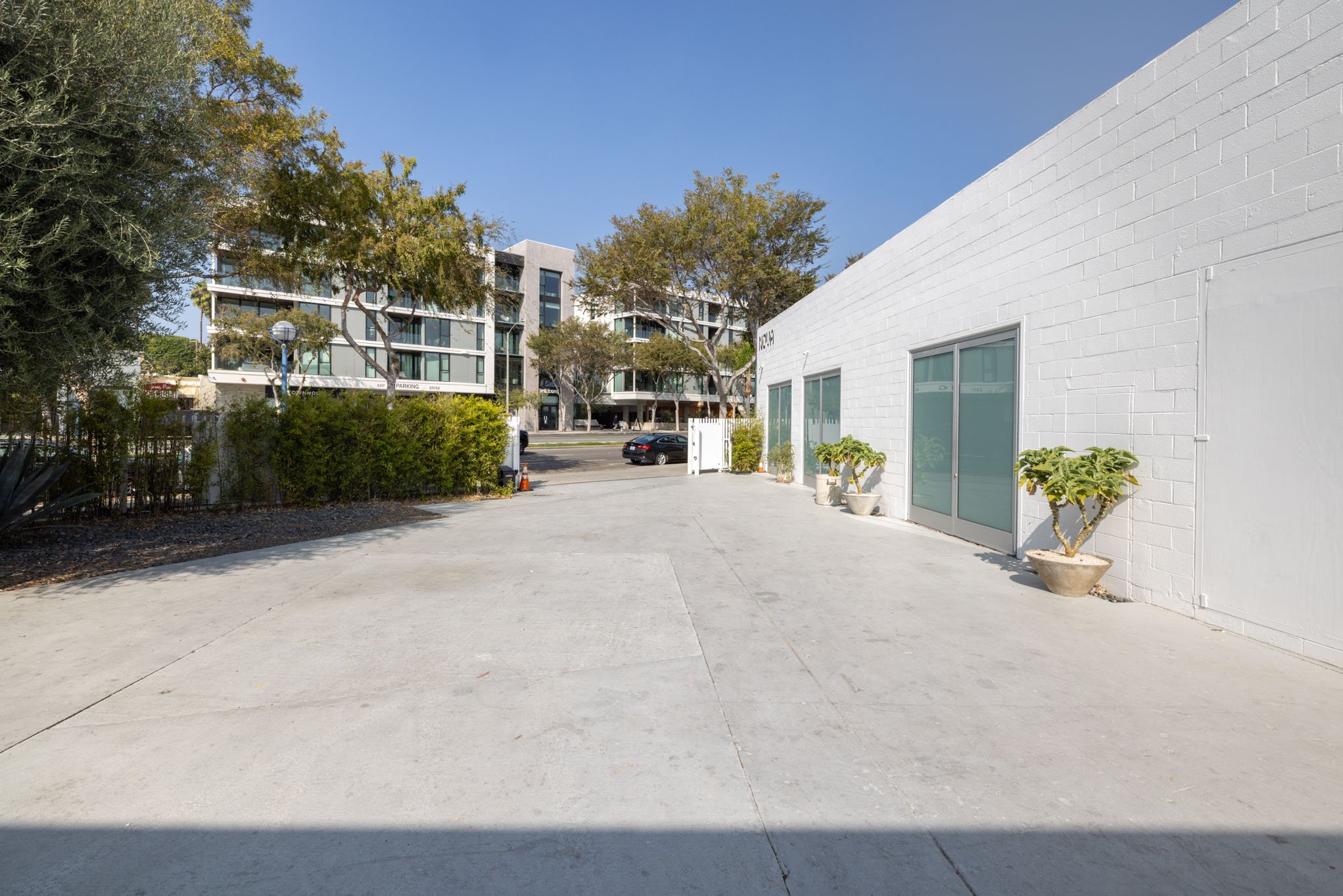 Empty parking lot with a white building on the right, potted plants, and a modern apartment complex in the background under a clear blue sky.