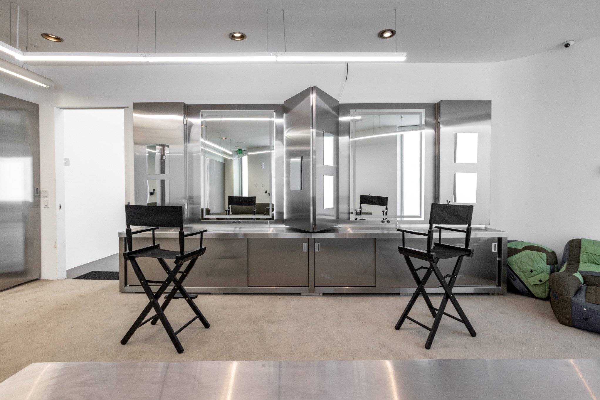 Modern salon station with large mirrors, black director chairs, and stainless steel cabinetry in a bright, minimalist space.