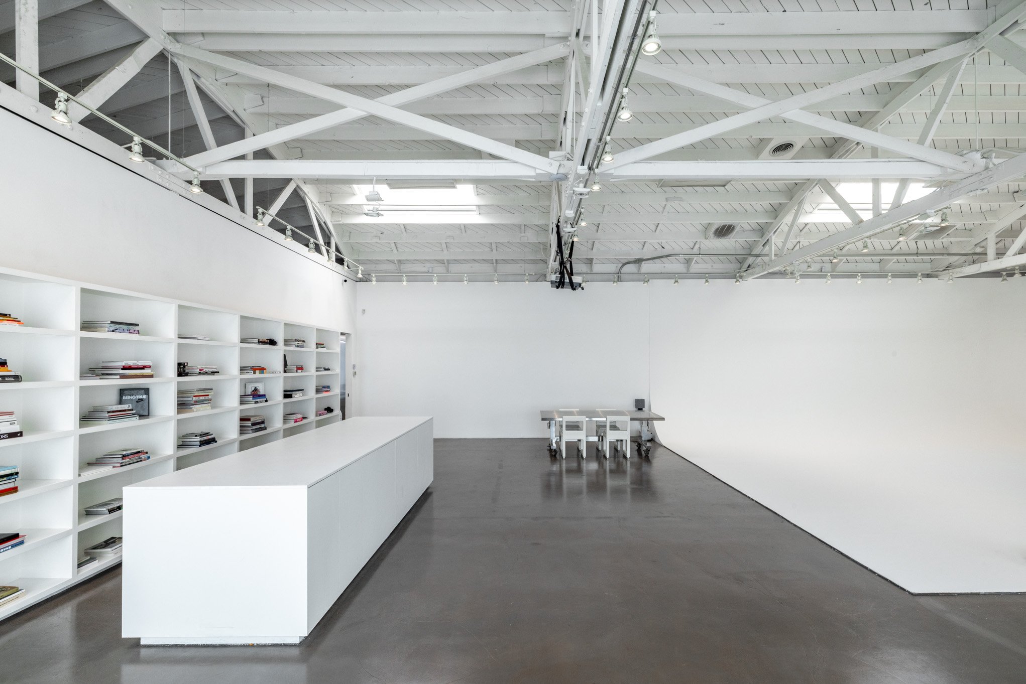 An empty art gallery or exhibition space with white walls, a white bookshelf filled with magazines or books, a long white table, and a small set of tables and chairs against the back wall. The ceiling has exposed white beams and track lighting.