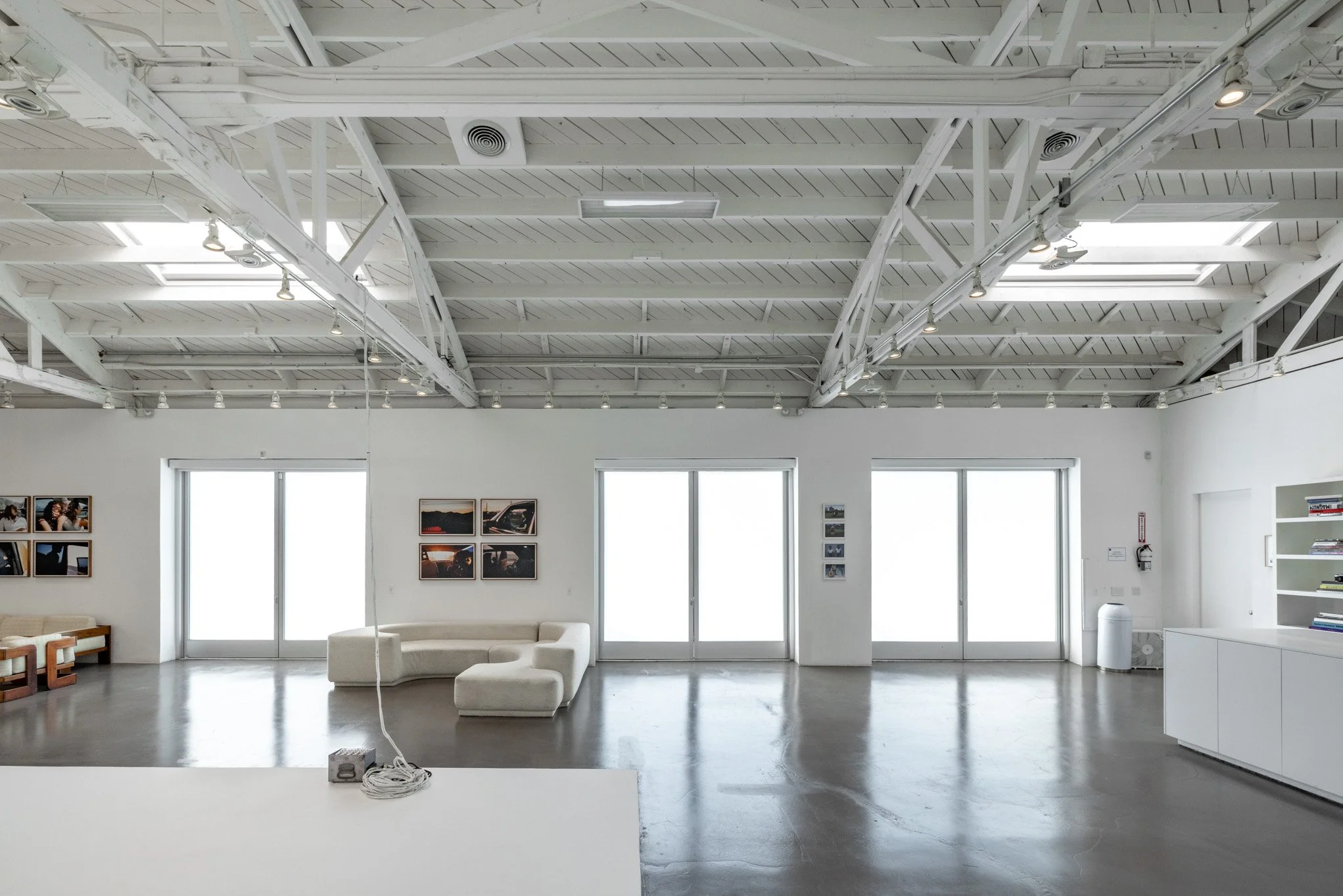 An art gallery with white walls, large windows, and a minimalist seating area with a white sectional sofa. The gallery features framed photographs on the walls and spotlights on the ceiling.