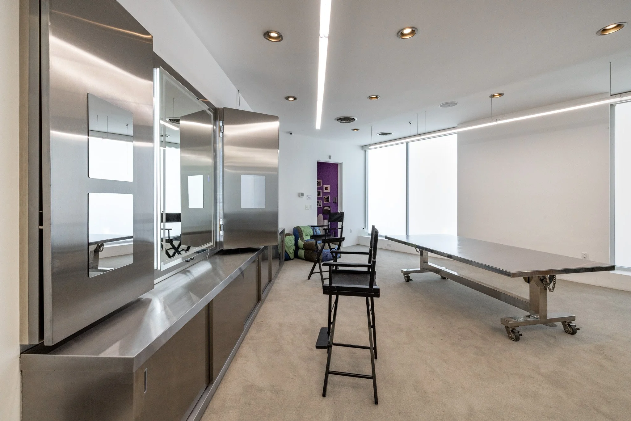 Modern office or conference room with stainless steel cabinets, chairs, and a large table on wheels, large windows with blinds, and a purple accent wall.