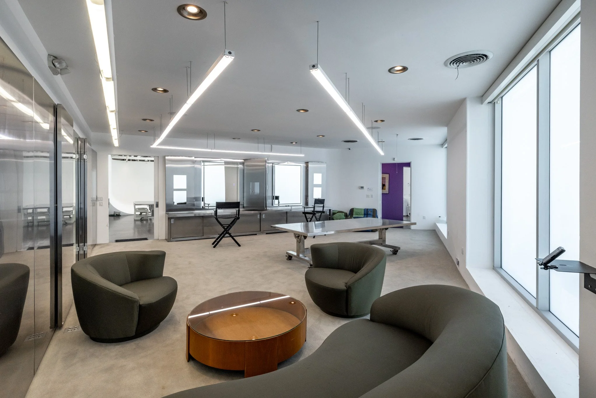 Modern office lounge area with green armchairs, a round wooden coffee table, large windows, and a white ceiling with linear lighting fixtures.
