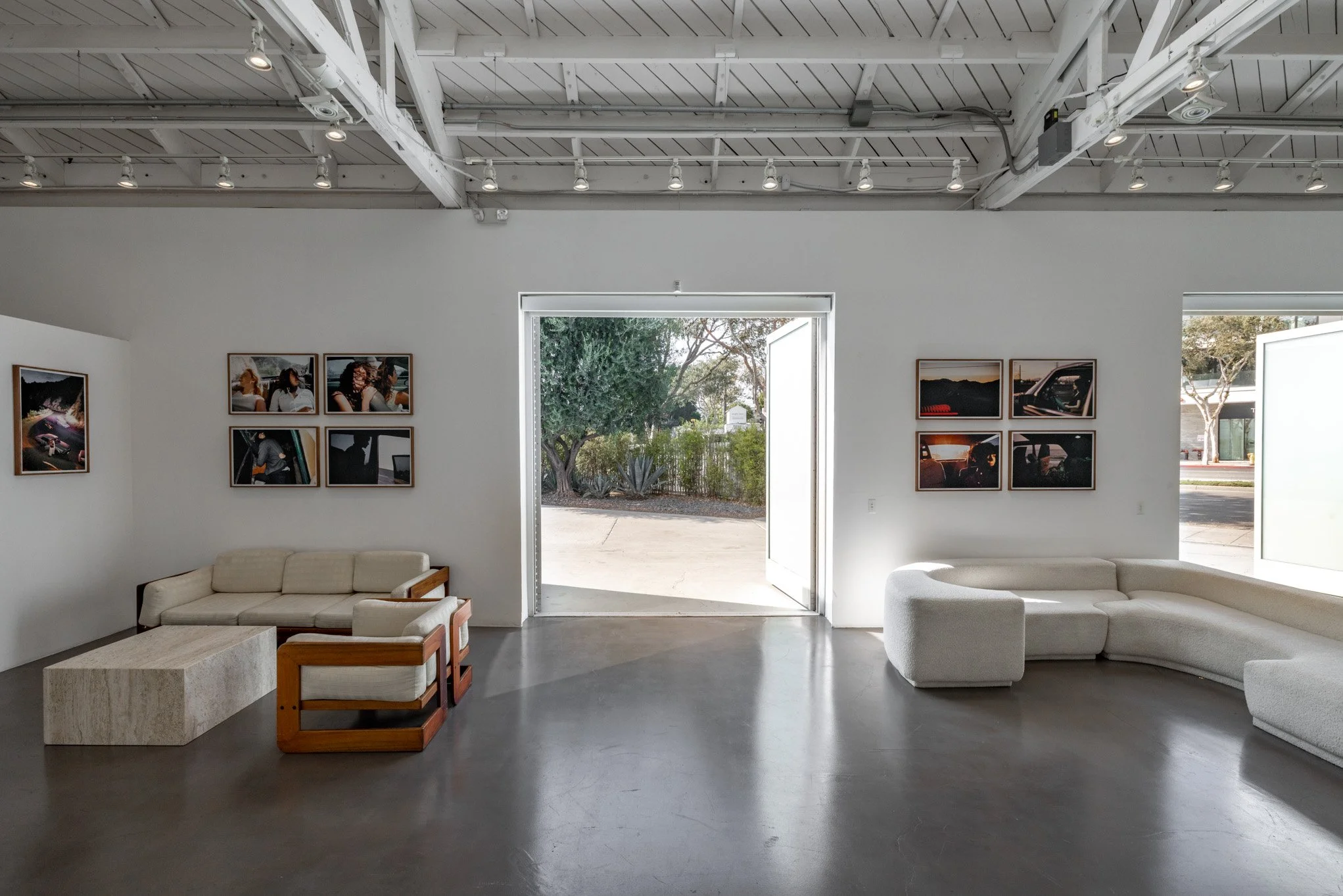 An art gallery with white walls, some framed photographs on the walls, modern white and wooden furniture, and an open door showing outdoor trees and sidewalk.