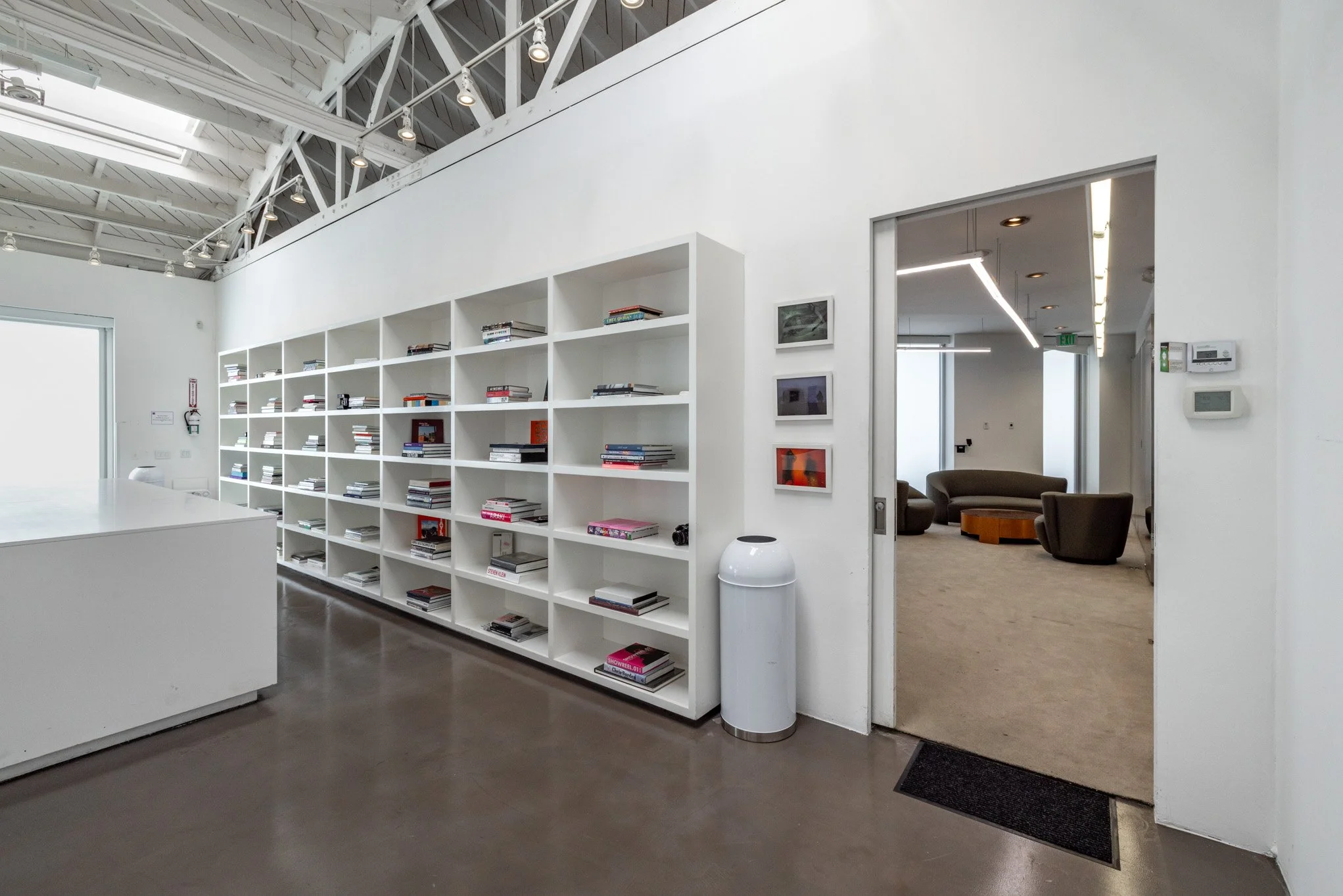 Interior of a modern office or lounge space with white walls, a large bookshelf filled with books, a doorway leading to a seating area with chairs and a rug, and abstract artwork on the walls.