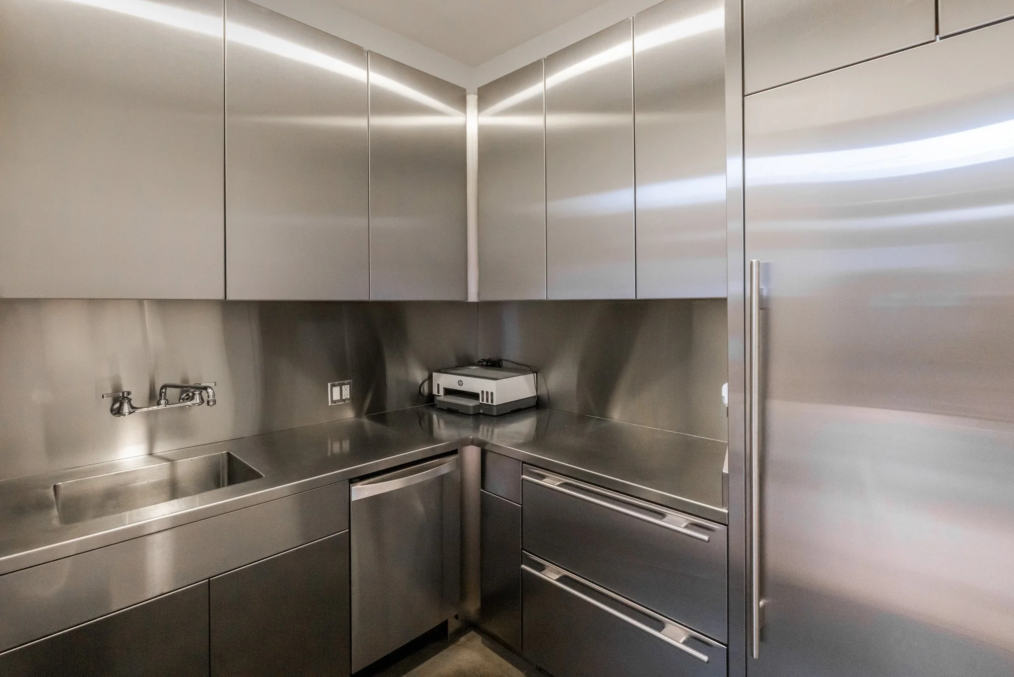 A modern, minimalist kitchen with stainless steel cabinets, sink, and countertop. There is a small appliance on the counter and a tall cabinet or refrigerator on the right.