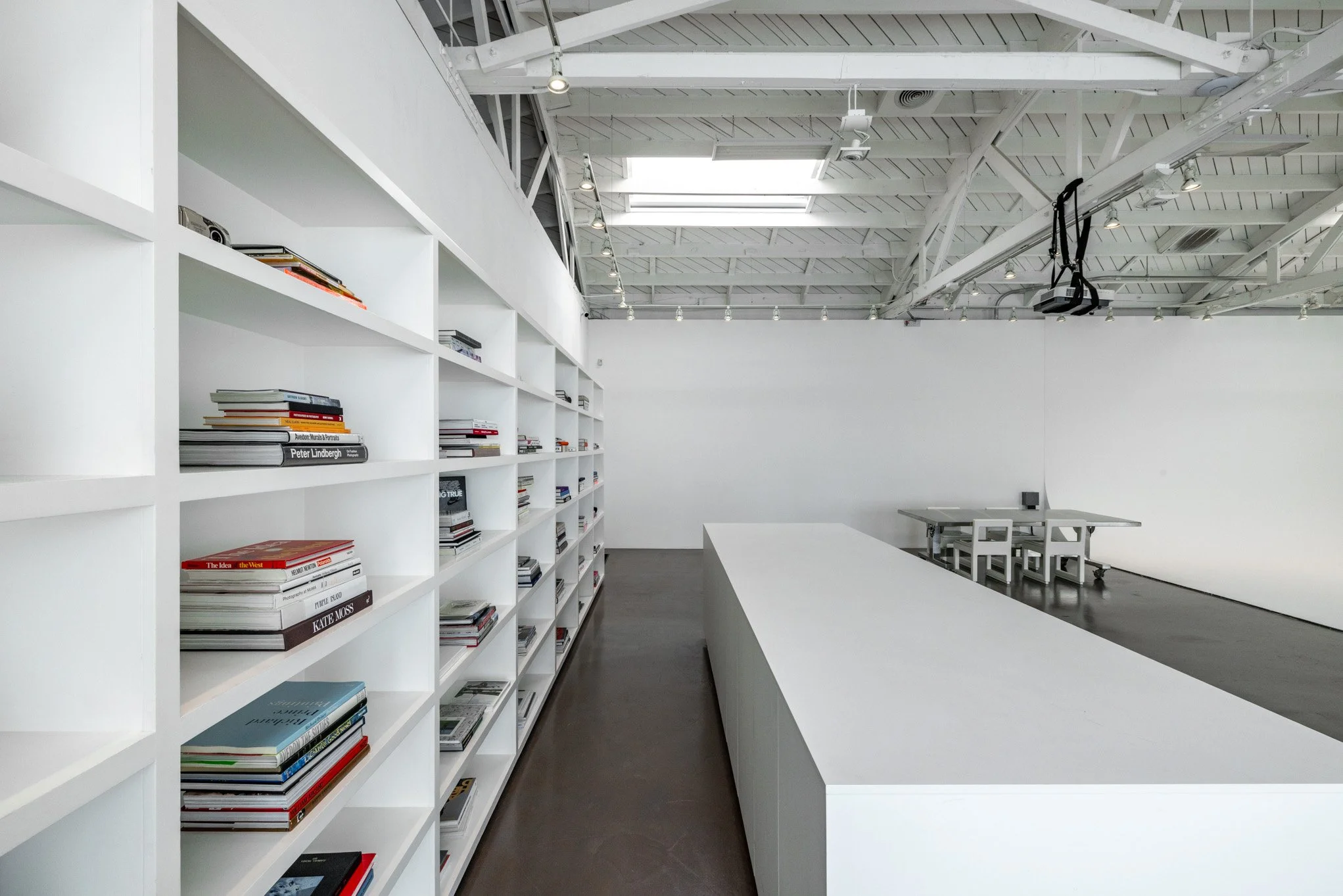 Empty modern art gallery with white walls, a long white table, and white chairs. White shelving filled with books on the left side and a ceiling with skylights and spotlights.