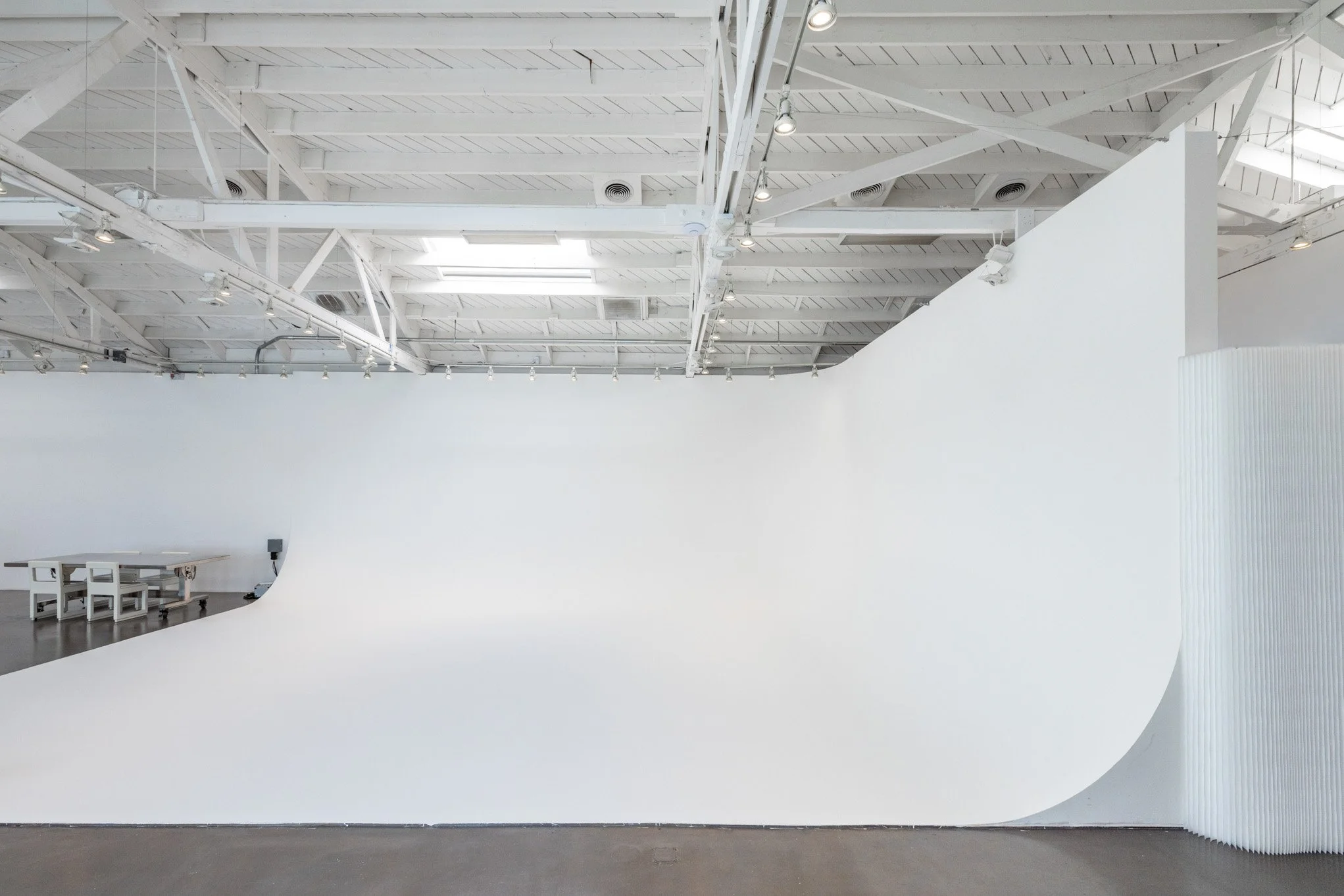 A large, white, curved backdrop in an art studio or gallery, with exposed white ceiling beams and lighting fixtures.