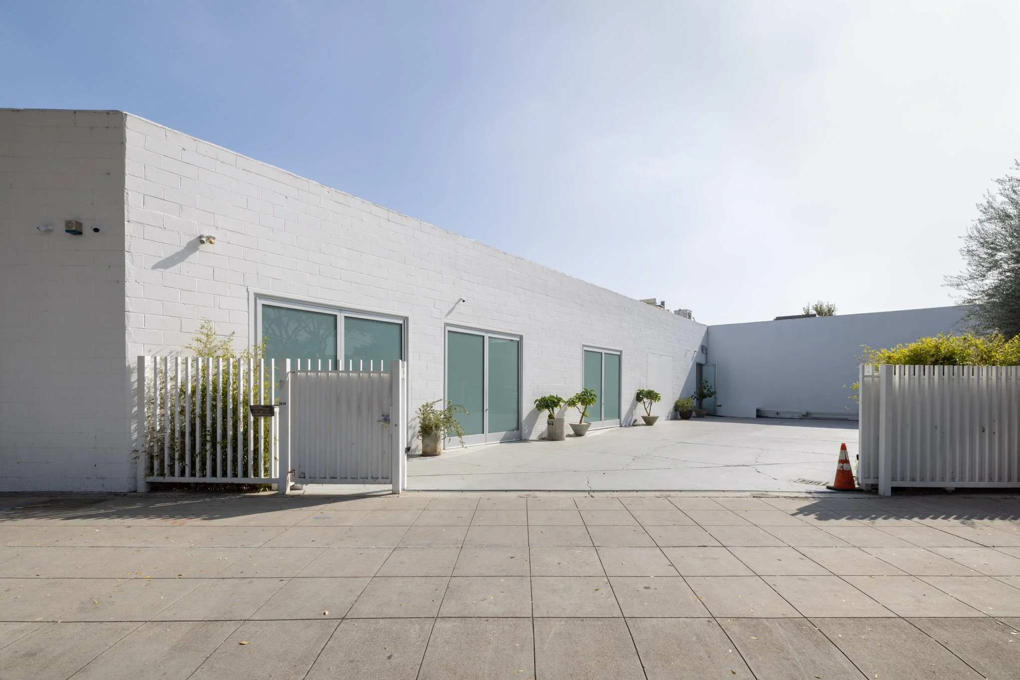 A modern white building with large glass sliding doors, potted plants outside, a white gate, and a tall white fence. The sky is clear and blue.