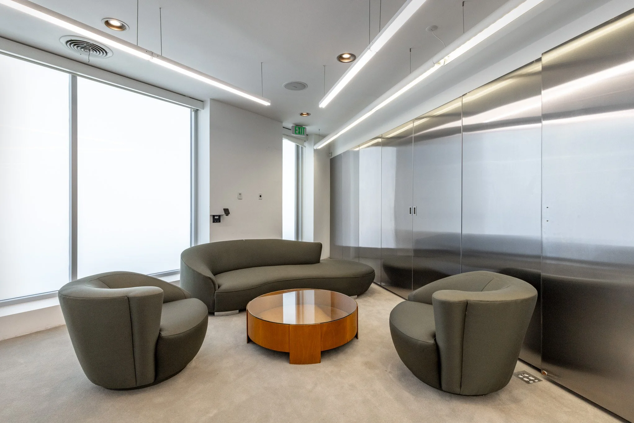 Modern interior lounge area with two rounded armchairs, a curved sofa, and a wooden coffee table, with large frosted windows and sleek metallic wall panels.