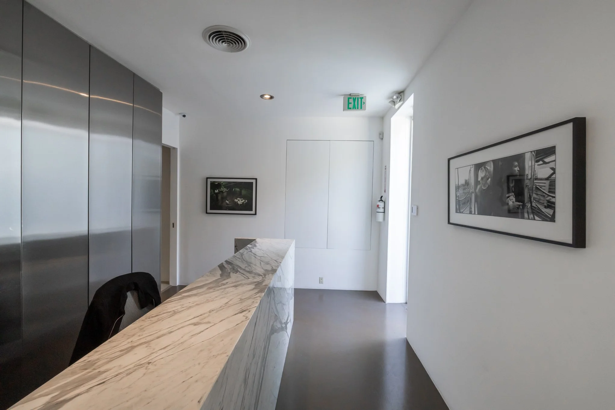 Modern reception area with marble countertop, black leather chair, framed black and white photo on white wall, metal elevator doors, and exit sign.