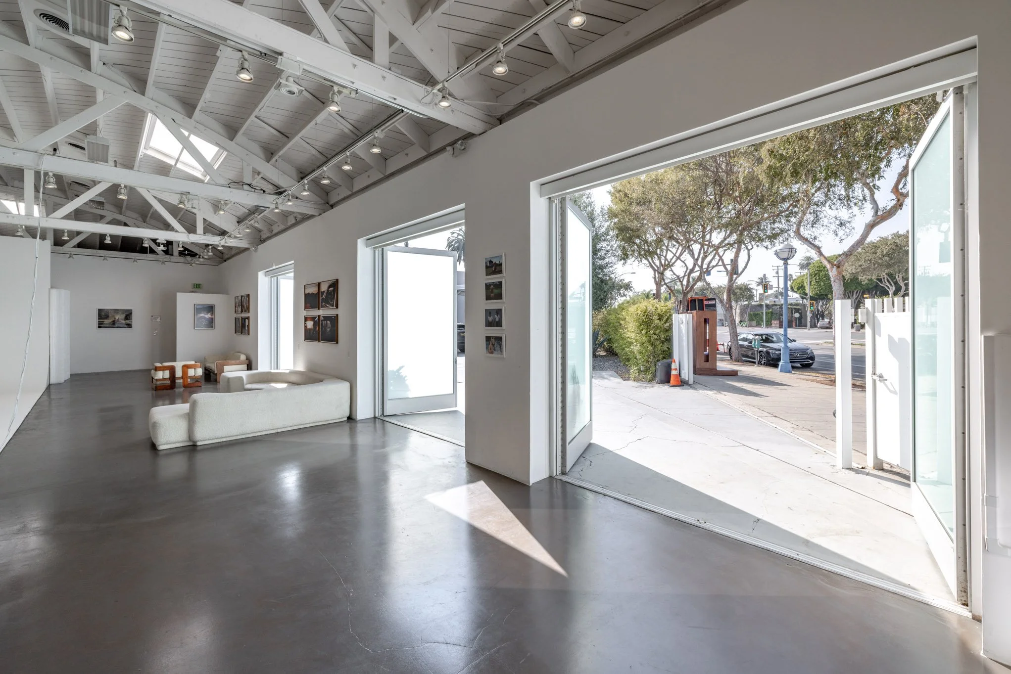 Interior of an art gallery with white walls, polished concrete floor, and partial ceiling with exposed beams and track lighting. Large open doors lead outside to a street with trees, parked cars, and a sidewalk.