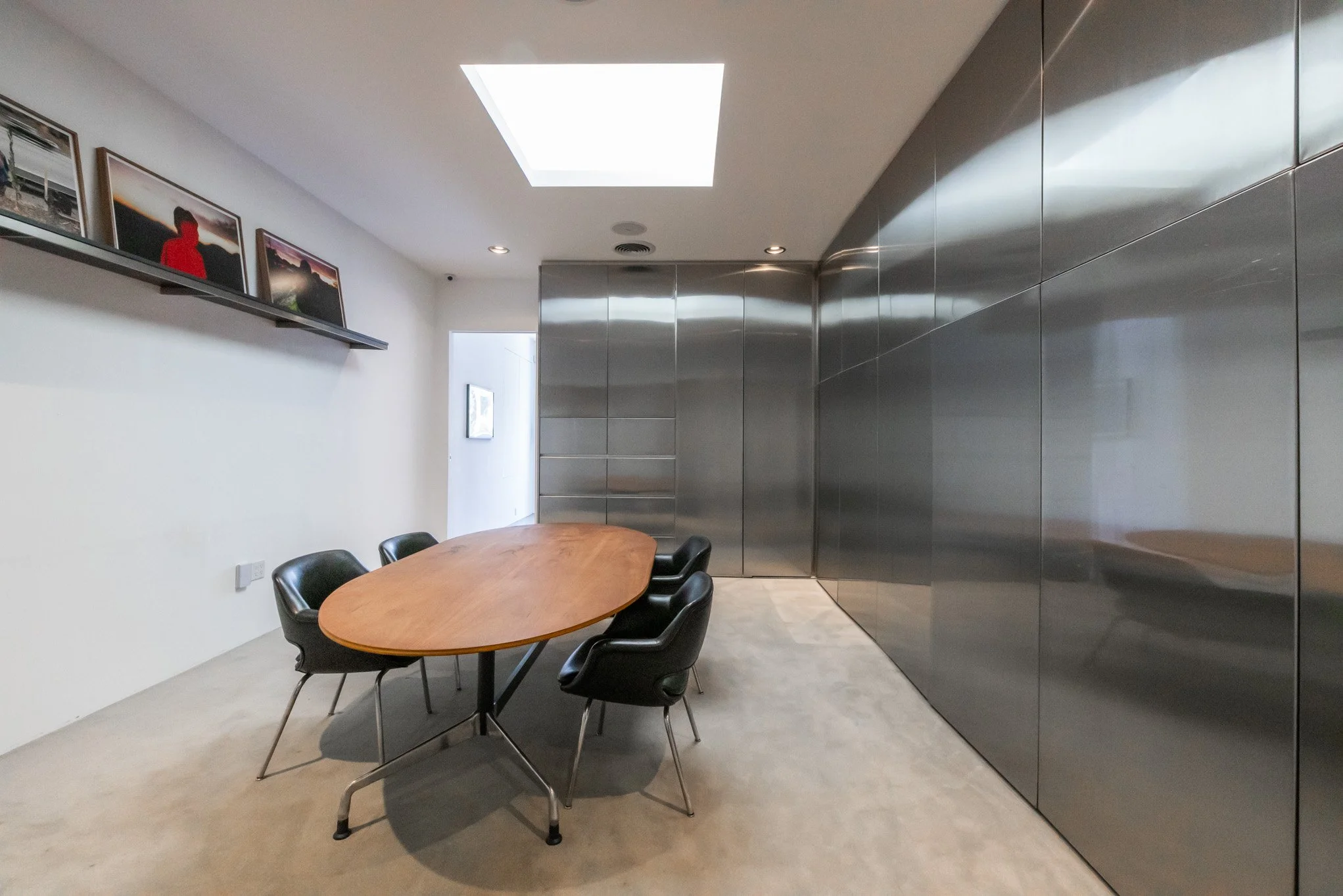 Modern conference room with a wooden oval table surrounded by six black chairs, a skylight on the ceiling, and a wall lined with large reflective metal panels. Artworks and photographs are displayed on a shelf and wall.