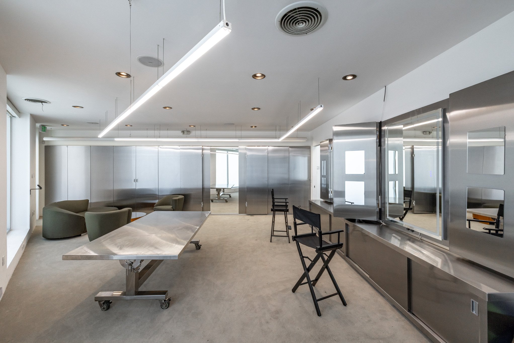 Modern office space with stainless steel countertops, chairs, and tables, illuminated by linear ceiling lights.