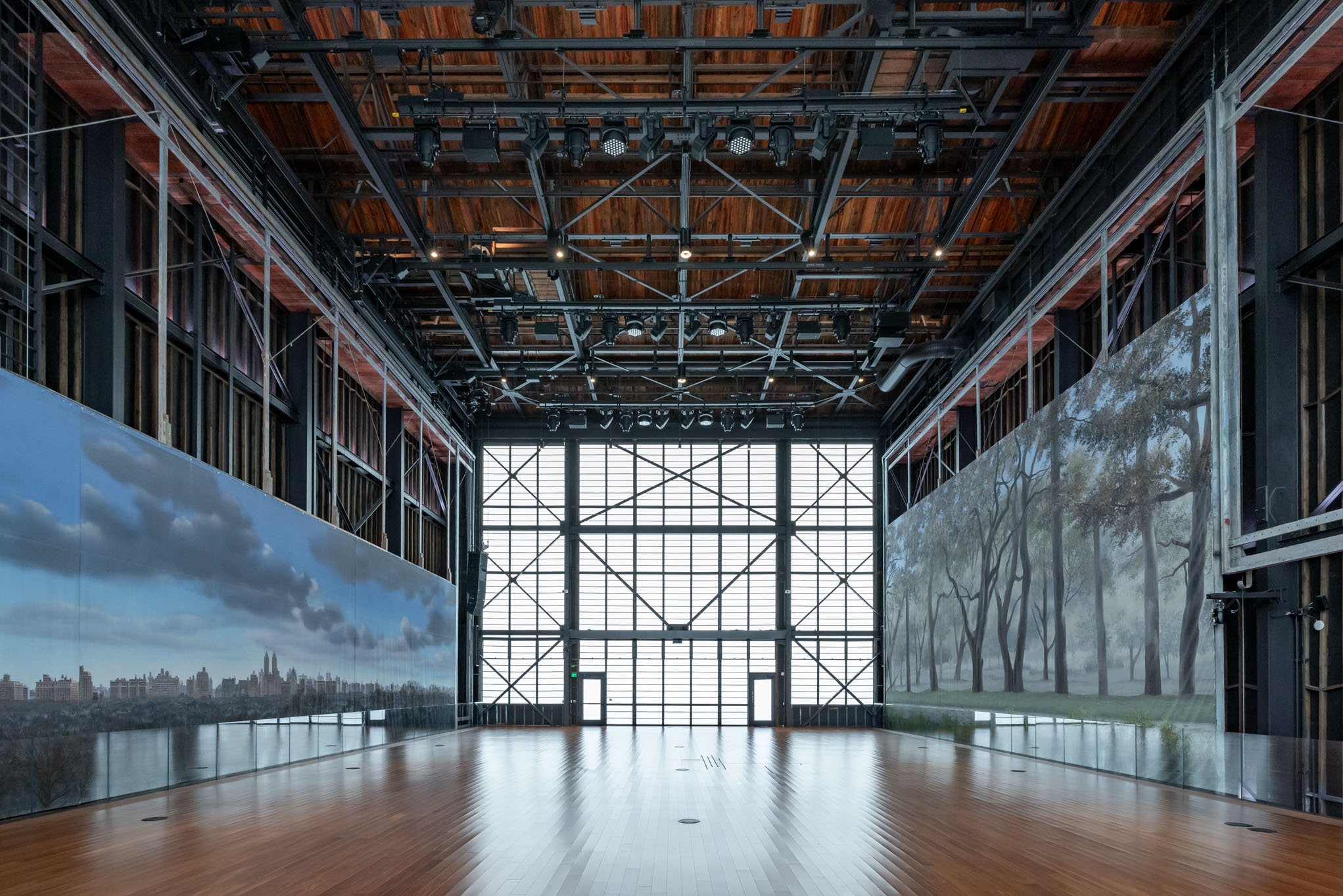 Empty art gallery or exhibition space with wooden flooring, large windows, artwork or murals of cityscape and trees on the walls, and a high ceiling with an industrial design.