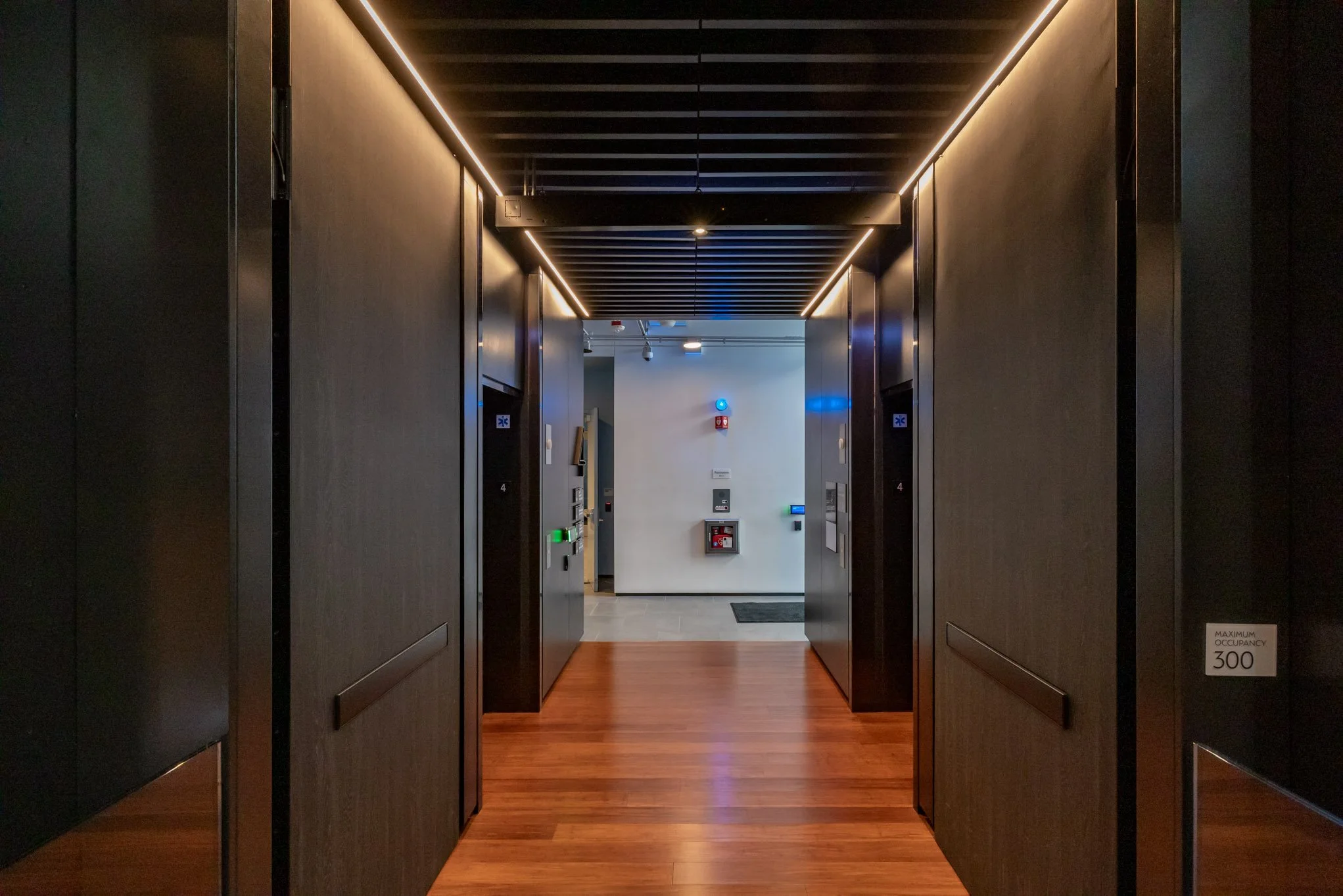 Inside an elevator lobby with dark walls, wooden flooring, and modern lighting, leading to elevator doors and a hallway with a fire alarm, emergency equipment, and a maximum occupancy sign.