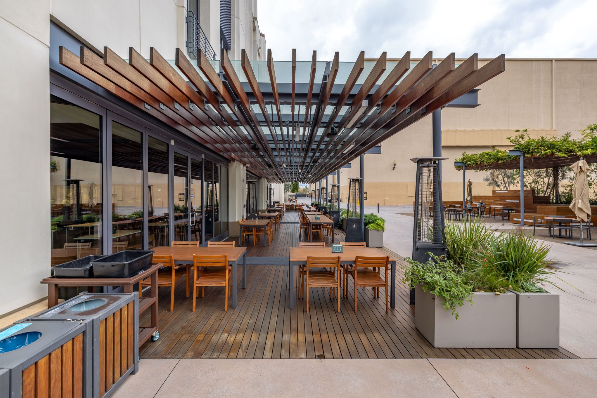 Empty outdoor restaurant patio with wooden tables, chairs, and heaters, covered by a wooden pergola, with some planters and a building with glass windows in the background.
