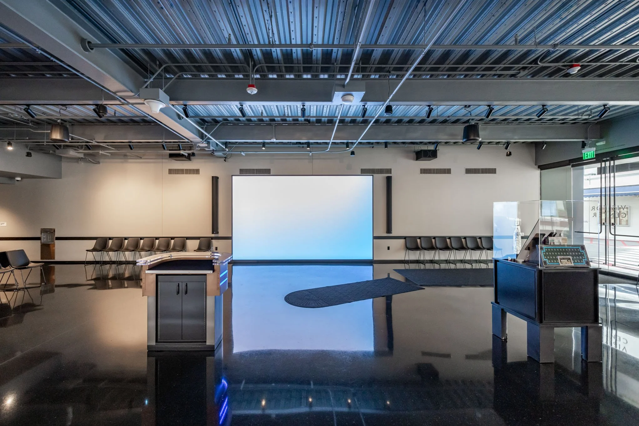 Empty conference or presentation room with a large screen at the front, black chairs lining the walls, and a small stage or podium in the center. Large windows on the right side with exit sign and glass door visible.