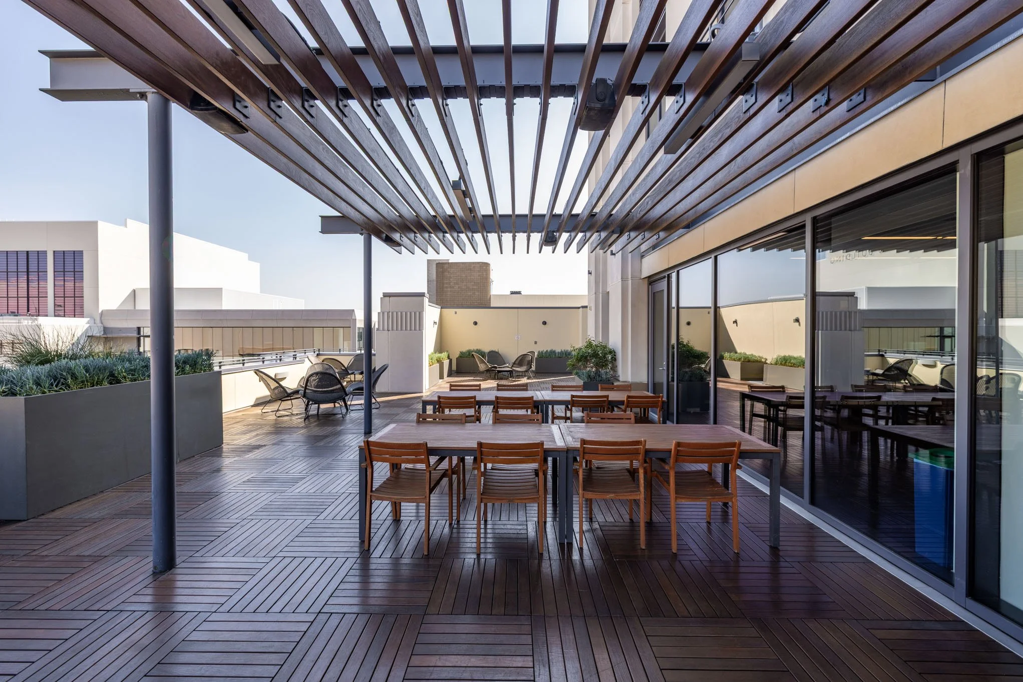 Empty rooftop terrace with wooden tables and chairs, potted plants, and a wooden slatted pergola.
