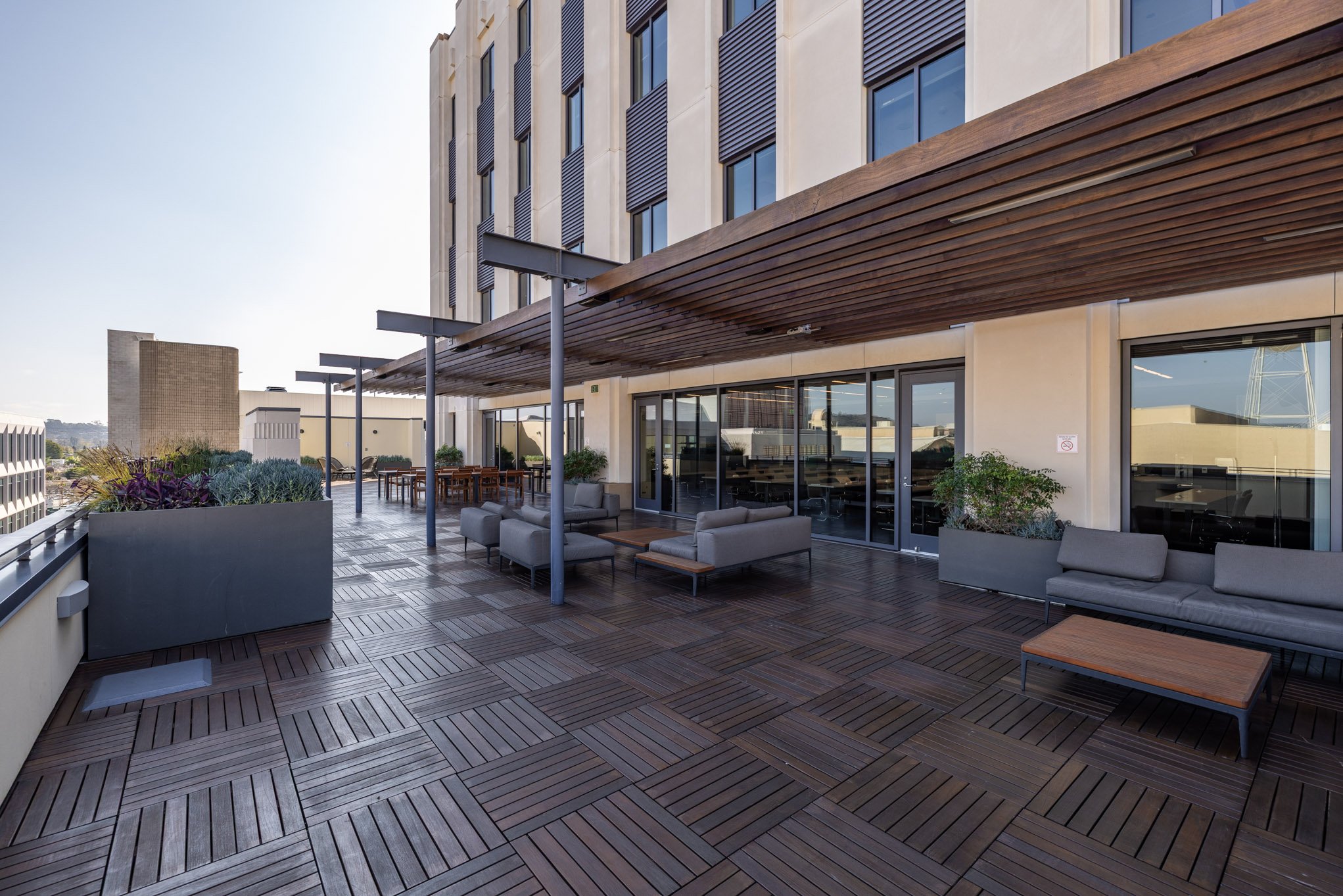 Empty outdoor rooftop terrace with wooden flooring, modern gray seating, potted plants, and city view in the background.