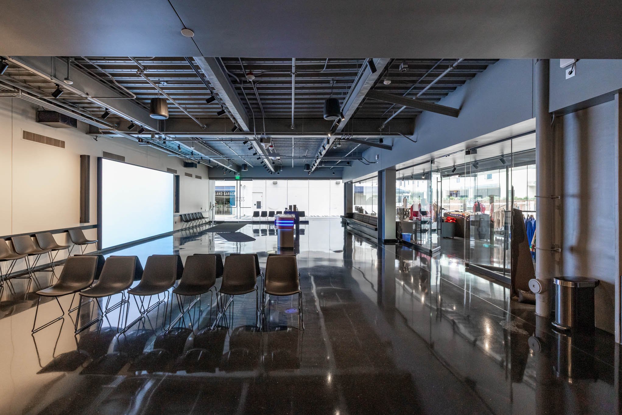 Empty waiting area with chairs and a large screen, glass wall separating from a retail space, and a shiny black floor in a modern building.