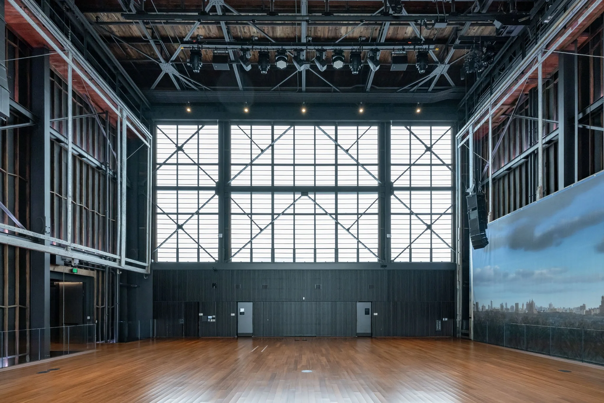 Empty modern indoor space with wooden flooring, large grid window, and black metal walls. Stage lighting and speakers are mounted on the ceiling.