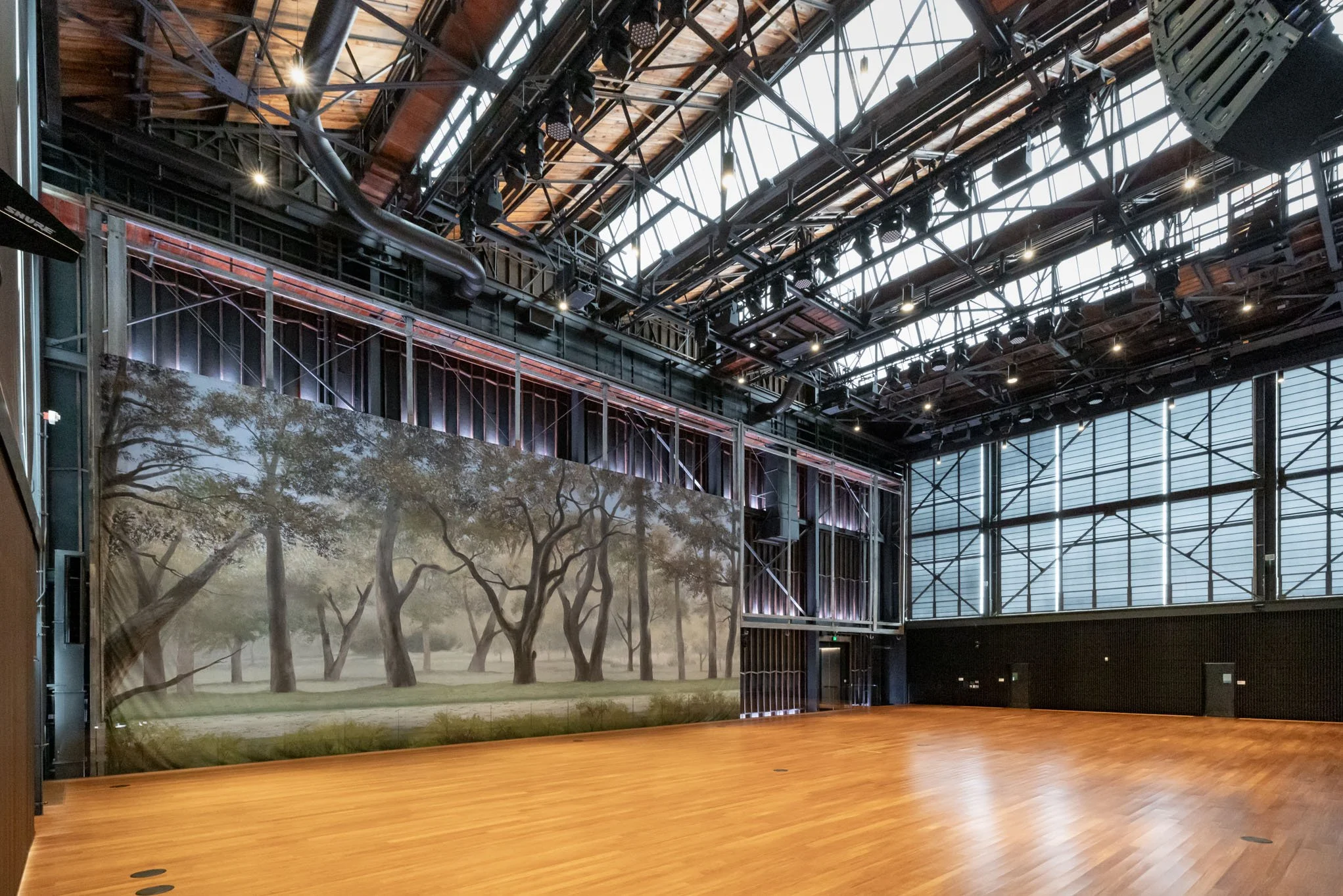 Empty indoor event space with wooden floor, large landscape painting of trees on one wall, black metal framework, and a high glass ceiling letting in natural light.