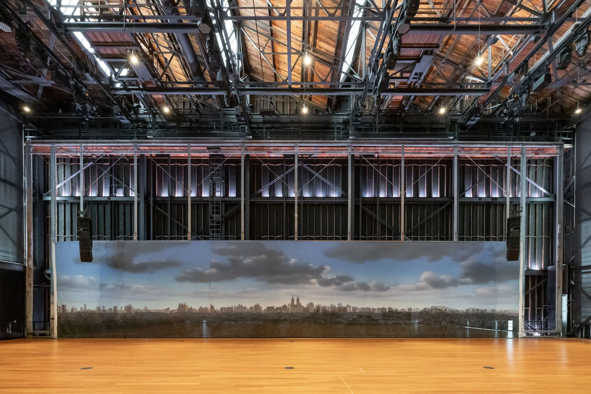 An indoor stage with a large panoramic backdrop of a city skyline under a partly cloudy sky, set against a wooden floor and metal framework ceiling.
