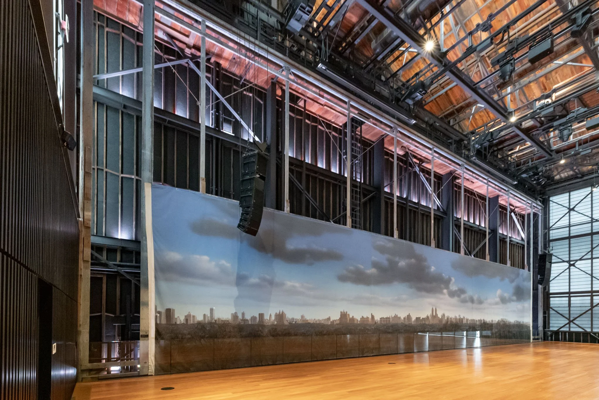 Interior of a large industrial-style space with wooden floors, a painted mural of a cityscape and sky, and visible metal framework and lighting fixtures overhead.