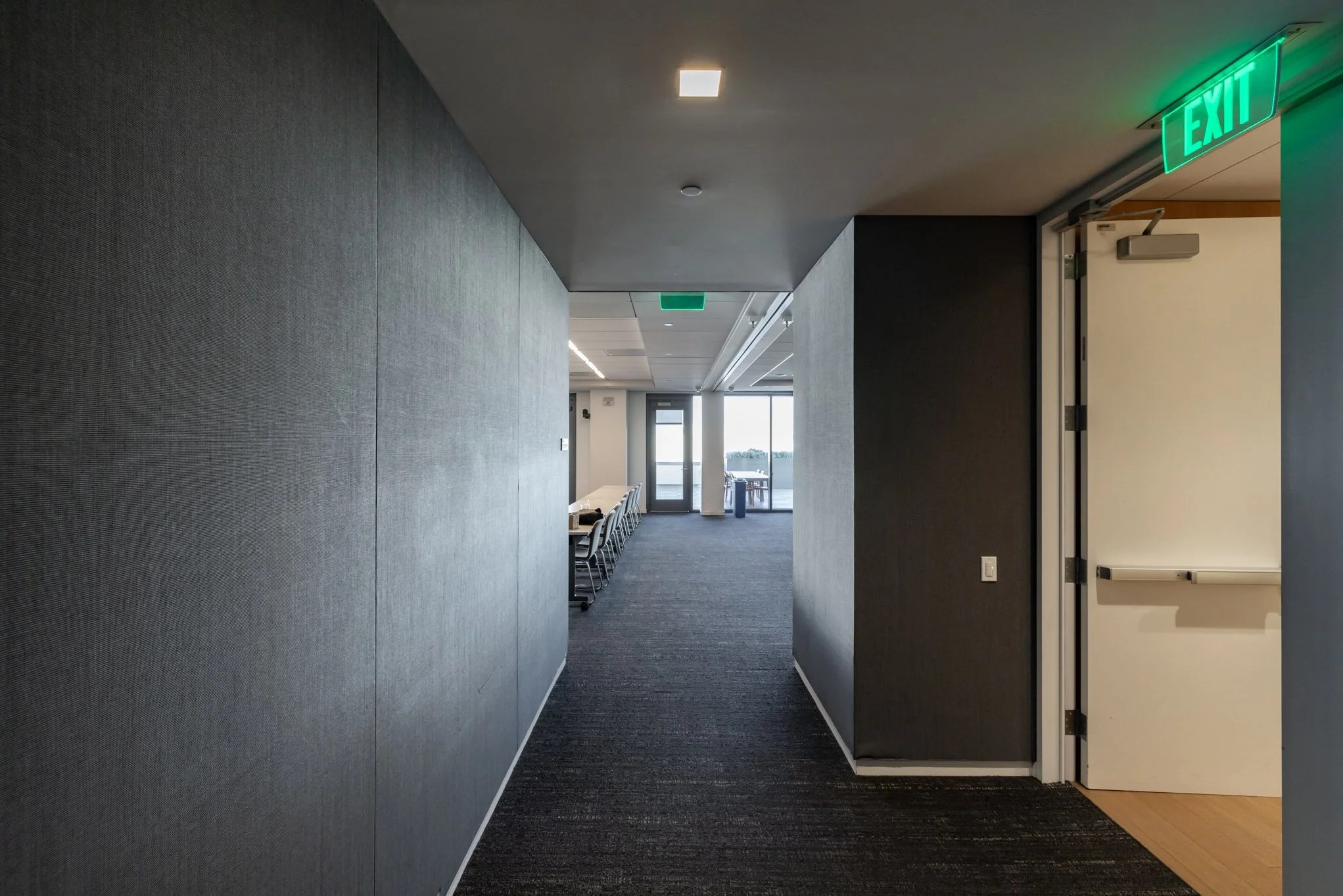 Corridor inside a modern office building with gray walls, carpeted floor, and green illuminated exit signs.