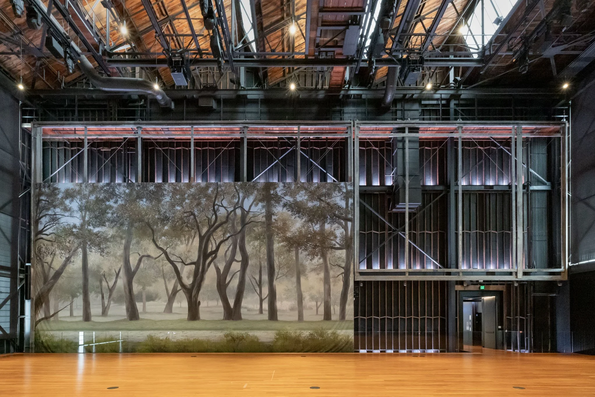 Empty stage with a large backdrop of a tree park scene, with the stage's metal framework and unfinished wall structure visible, in an indoor theater or studio space.