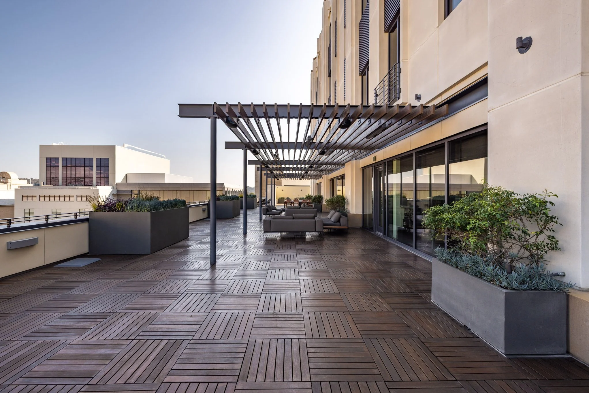 Large outdoor balcony with wooden deck flooring, modern potted plants, and outdoor seating including sofas and a dining table with chairs. A wooden pergola provides partial shade over the seating area, and glass sliding doors lead to the interior of 