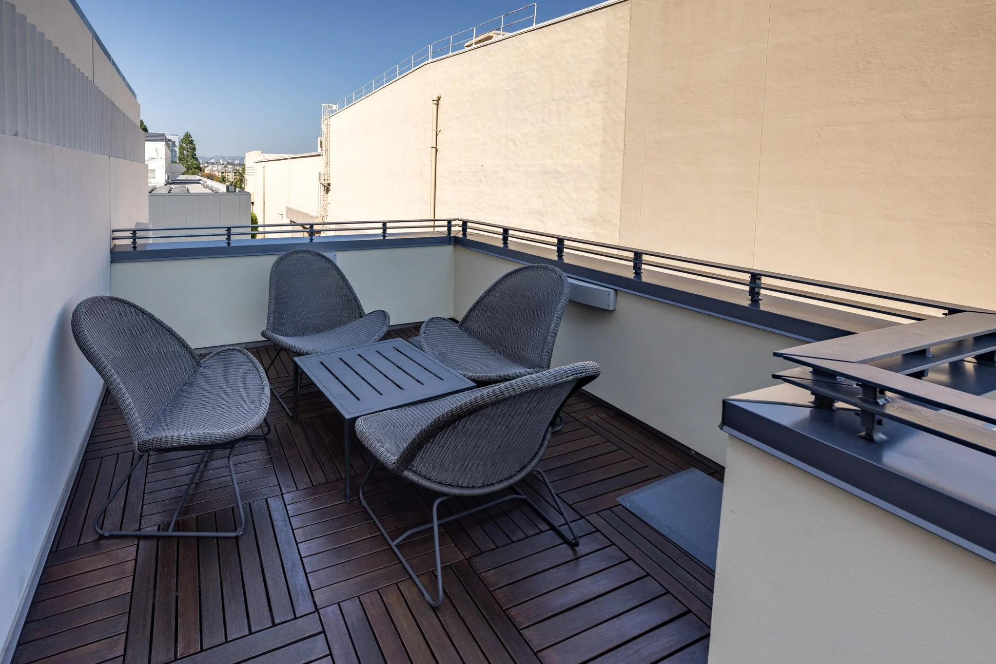 Small outdoor balcony with four gray woven chairs and a gray table on a wooden deck, overlooking neighboring buildings and a clear blue sky.