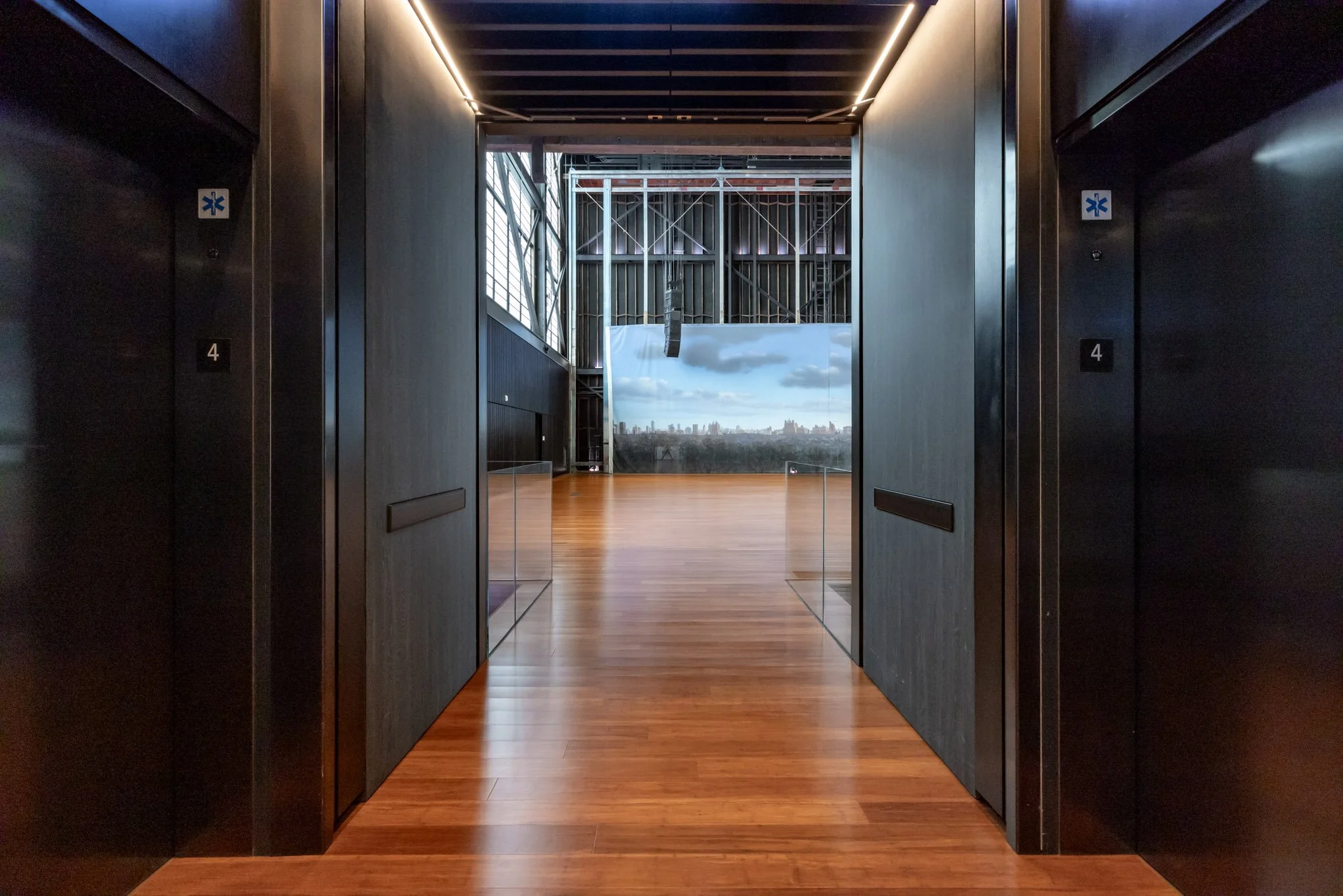 View through an elevator doorway into a spacious room with wooden floors and a large window showing a city skyline in the distance.