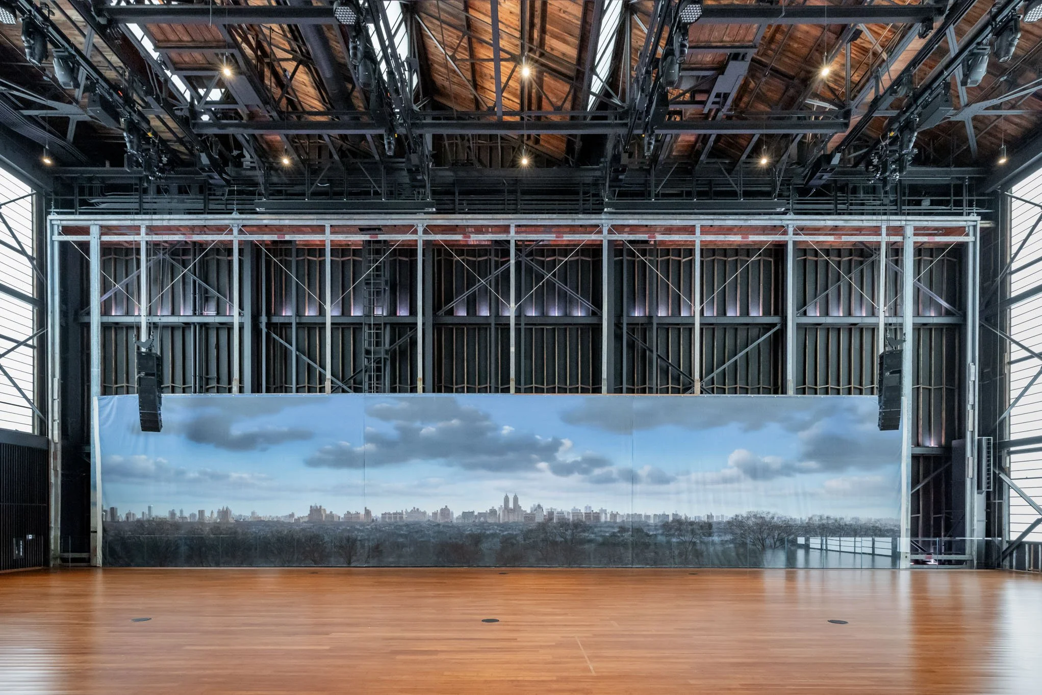 Empty indoor stage with a city skyline backdrop, wooden floor, and metal scaffolding on the walls.