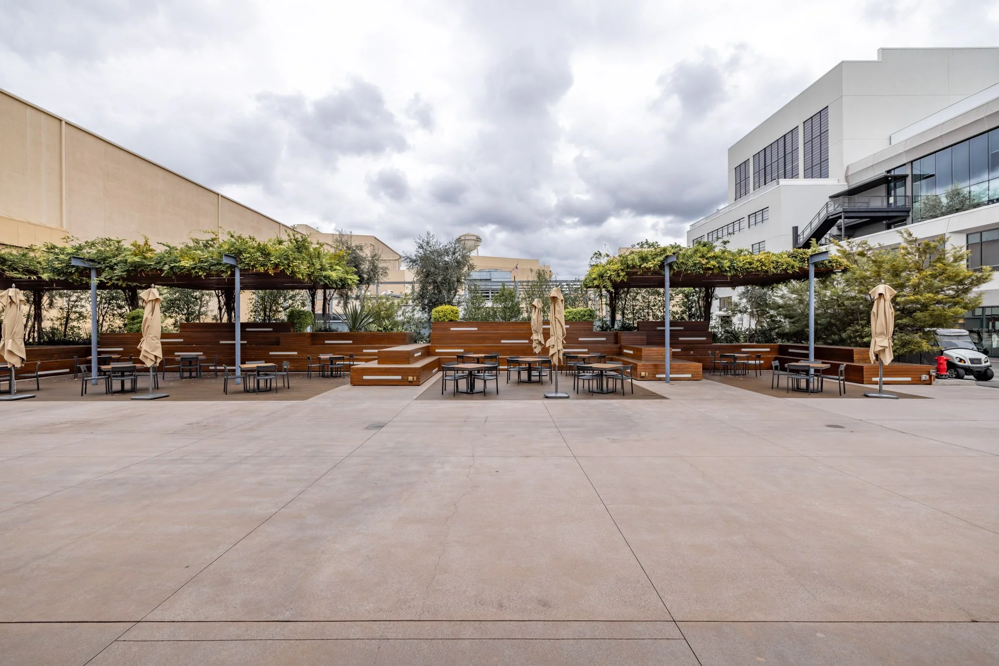 Empty outdoor terrace with seating and shaded areas, surrounded by modern buildings, under cloudy sky.