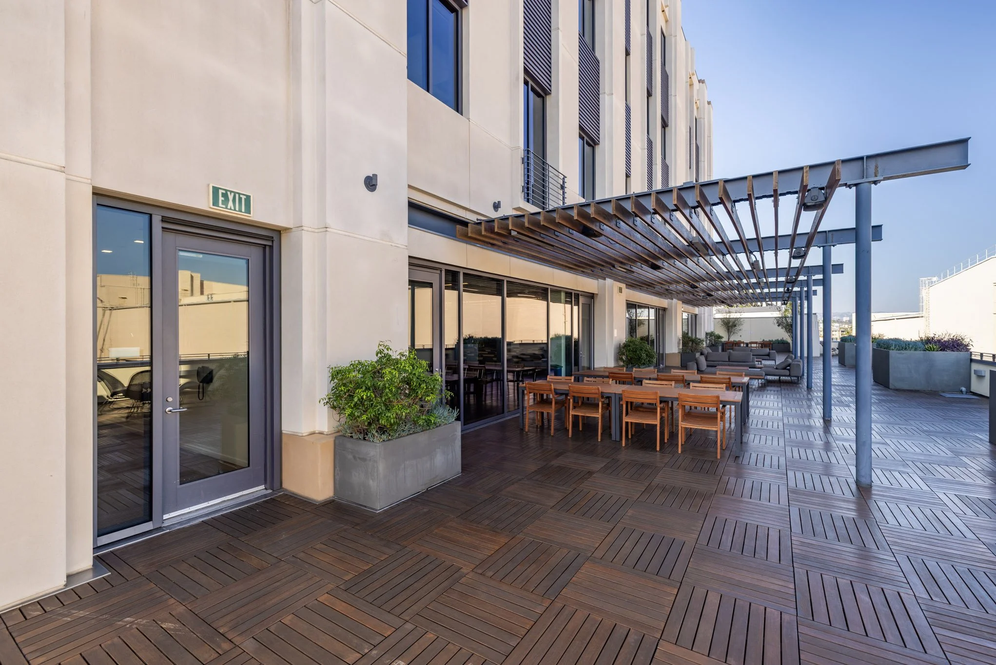 Empty outdoor rooftop patio with wooden flooring, wooden tables and chairs, gray sofas, potted plants, and a wooden pergola on a sunny day.