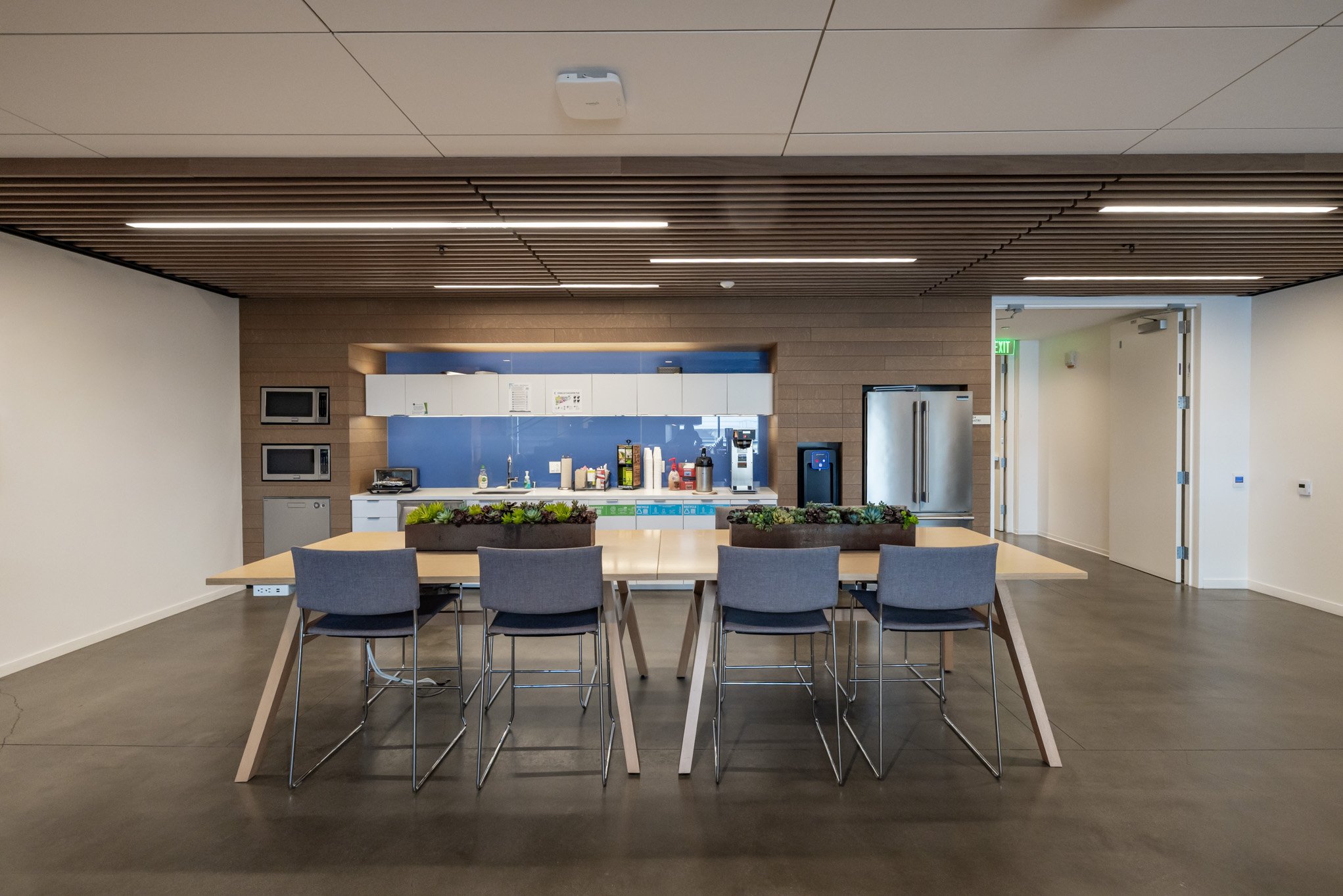 Modern office kitchenette with a long table, six chairs, plants in the center, and appliances including microwaves, a refrigerator, and a coffee machine.