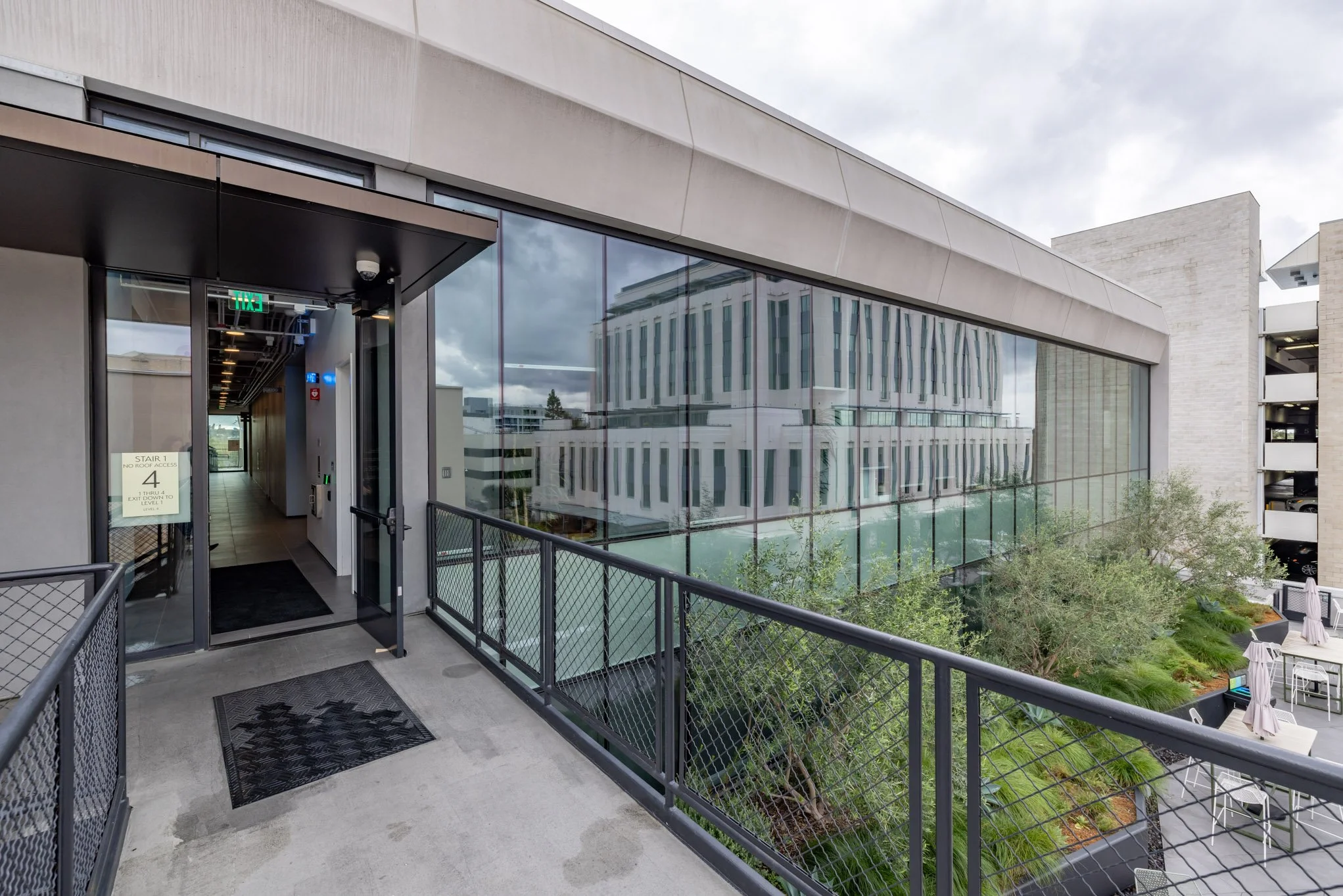Exterior view of a modern building with a glass facade and a balcony with a metal railing. The sky is cloudy, and there is outdoor seating with umbrellas on the patio below.