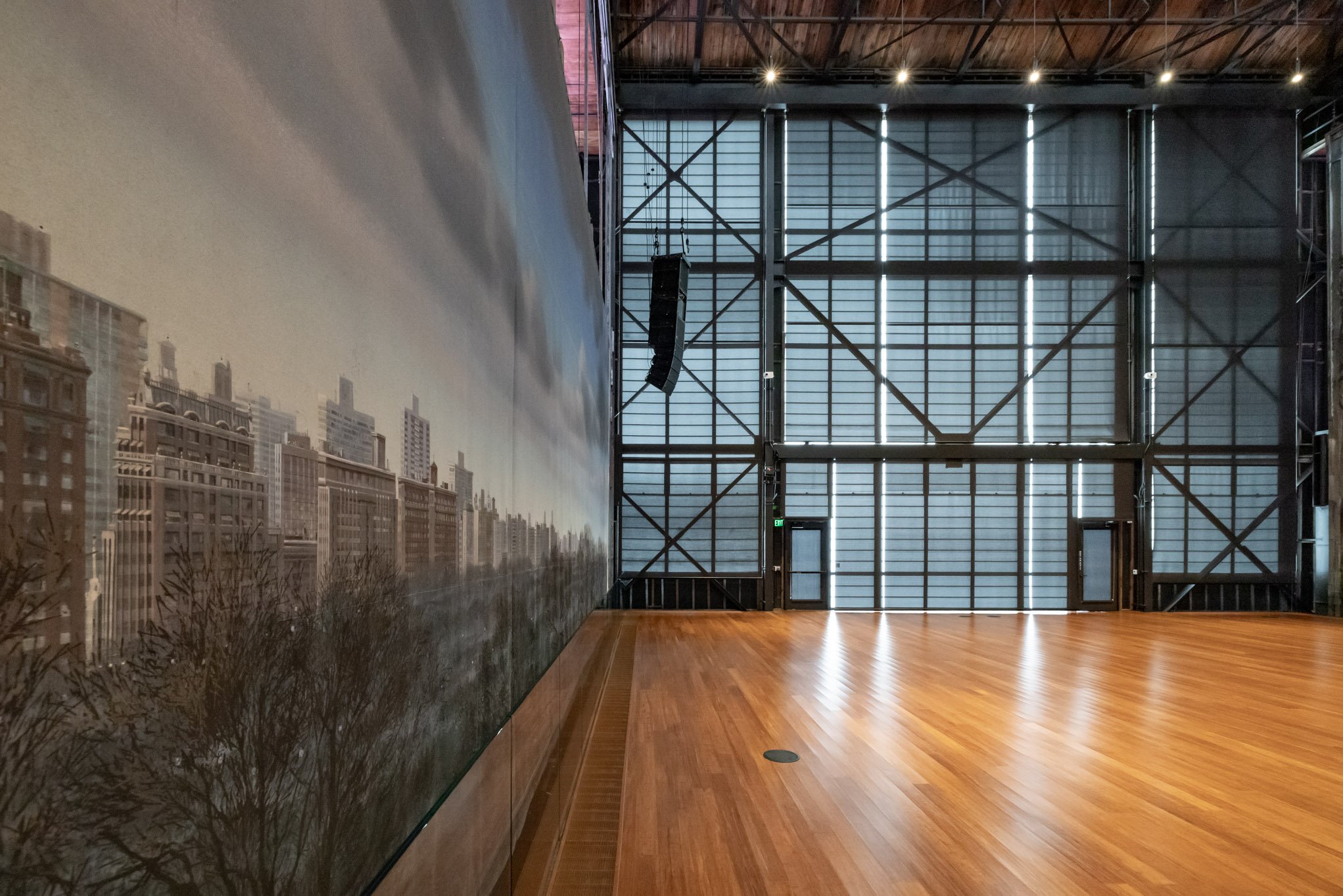 An empty room with a wooden floor, a large wall mural of a city skyline, and a high ceiling with metal framework, illuminated by ceiling lights.