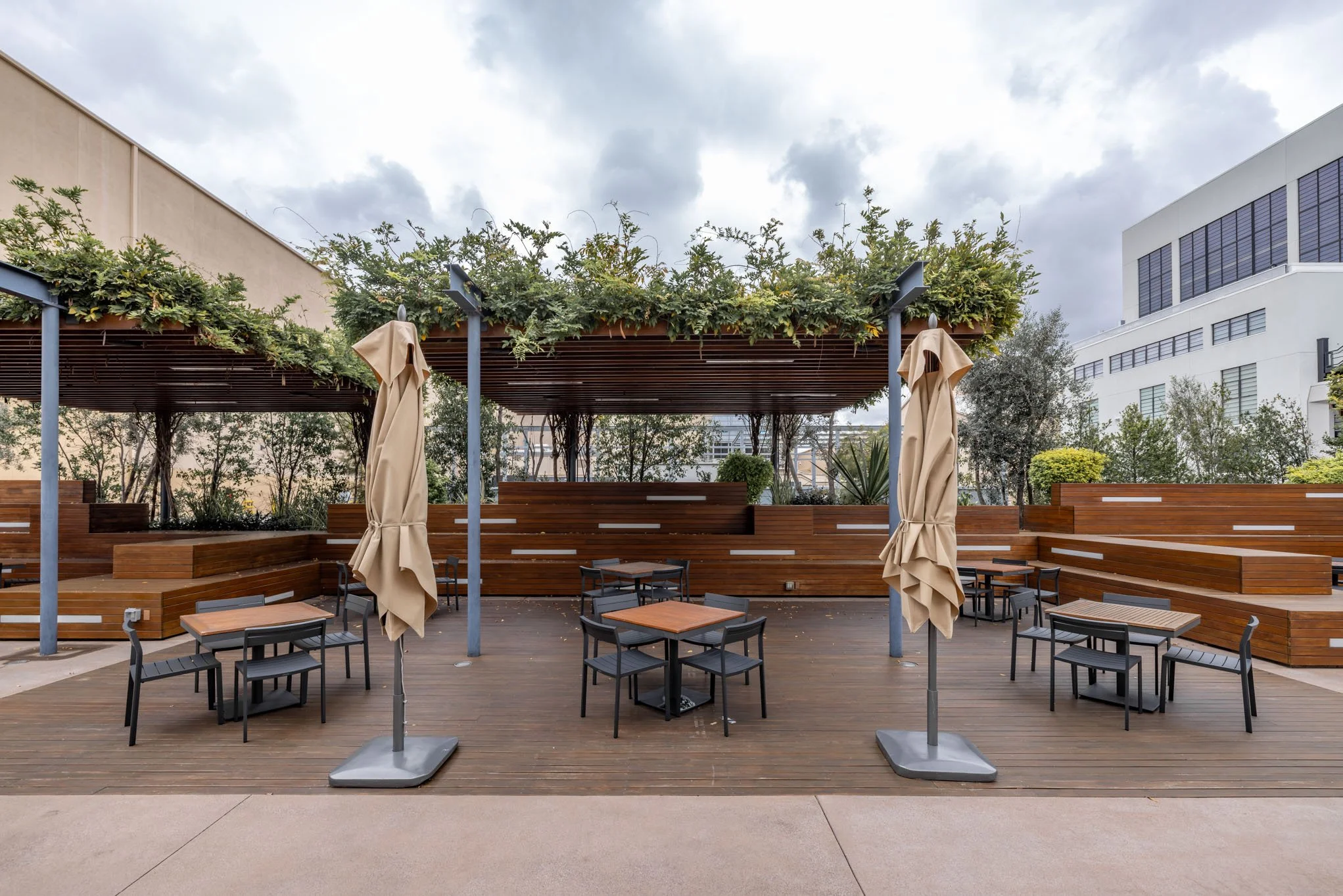 Empty outdoor patio with wooden decking, tables, black chairs, closed beige umbrellas, and a leafy pergola, with modern buildings and overcast sky in the background.