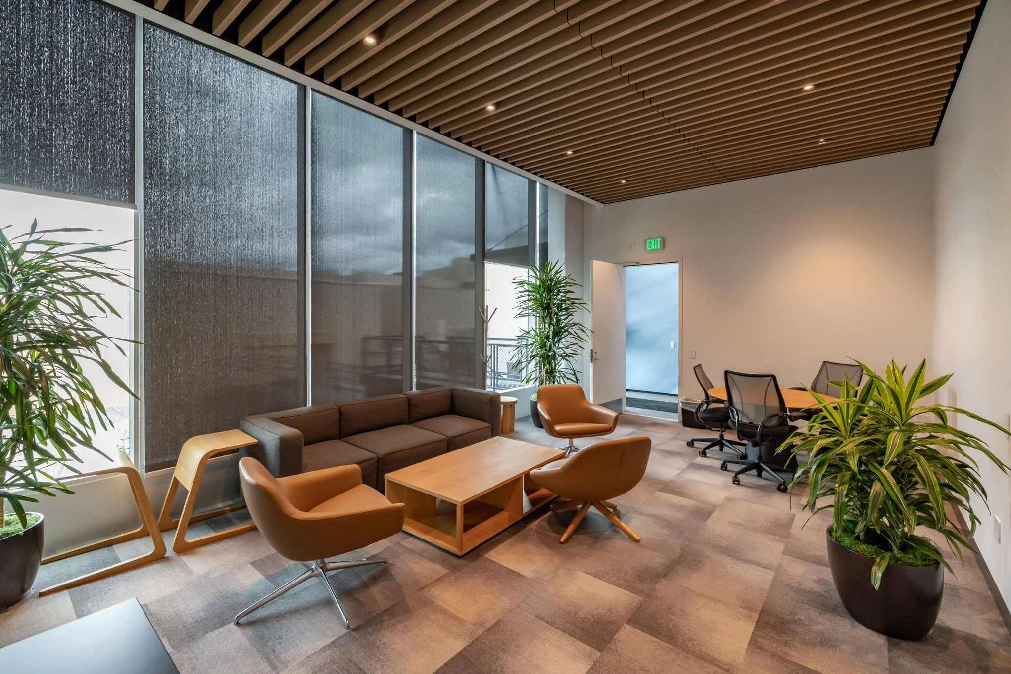 Modern office waiting area with brown and orange chairs, a brown sofa, wooden coffee table, potted plants, glass wall, and a door with a green exit sign.