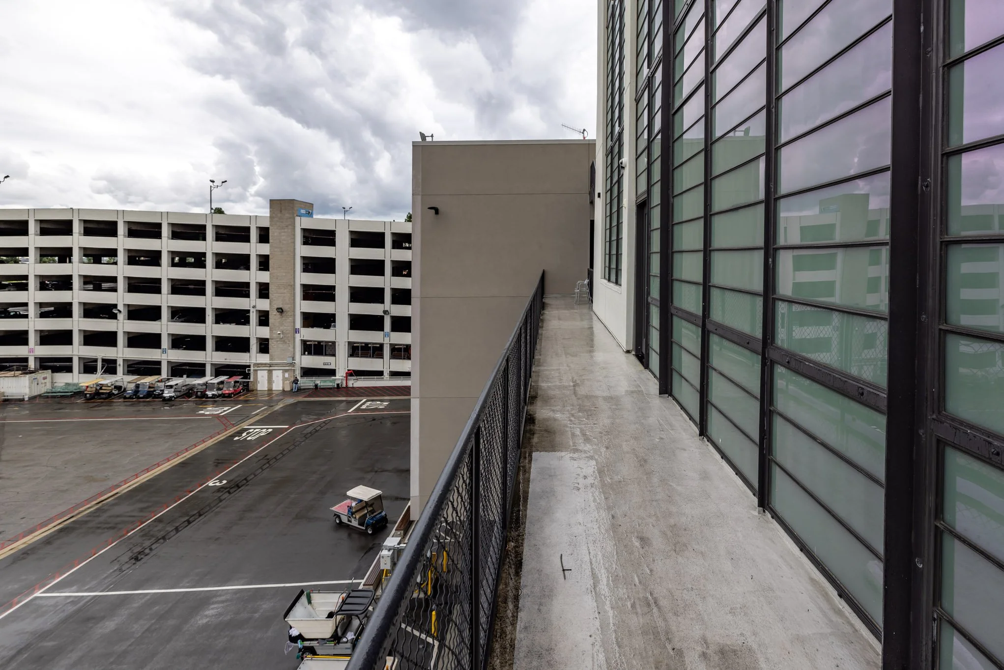 Empty outdoor balcony with a metal railing, concrete flooring, and a view of a parking garage and cloudy sky.