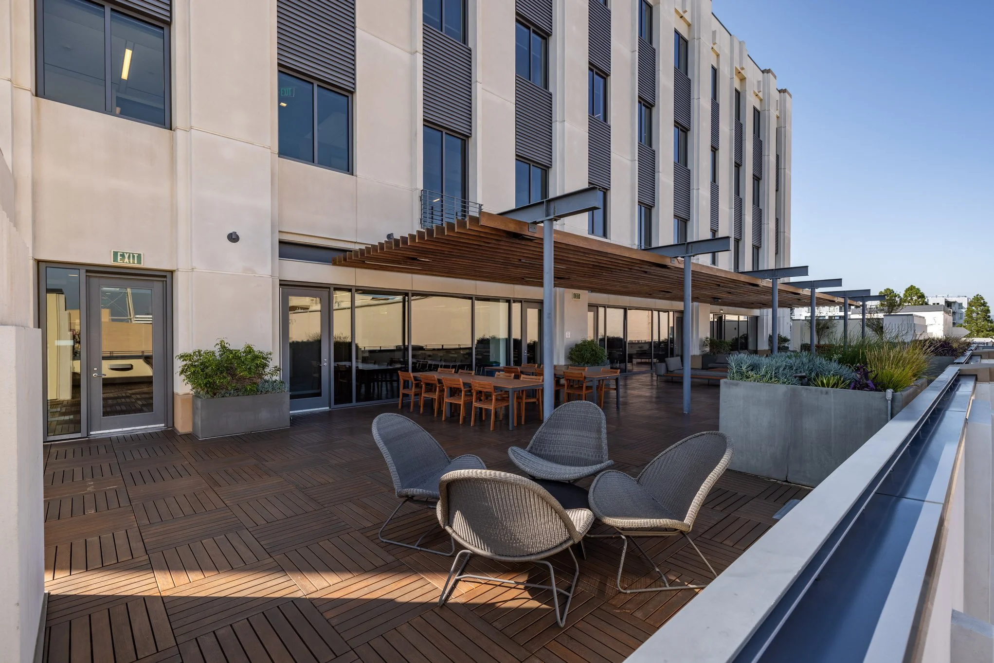 Outdoor terrace with wooden flooring, modern chairs arranged in a circle, and a dining table with chairs beneath a wooden pergola, against the backdrop of a contemporary building with large windows and planters with greenery.