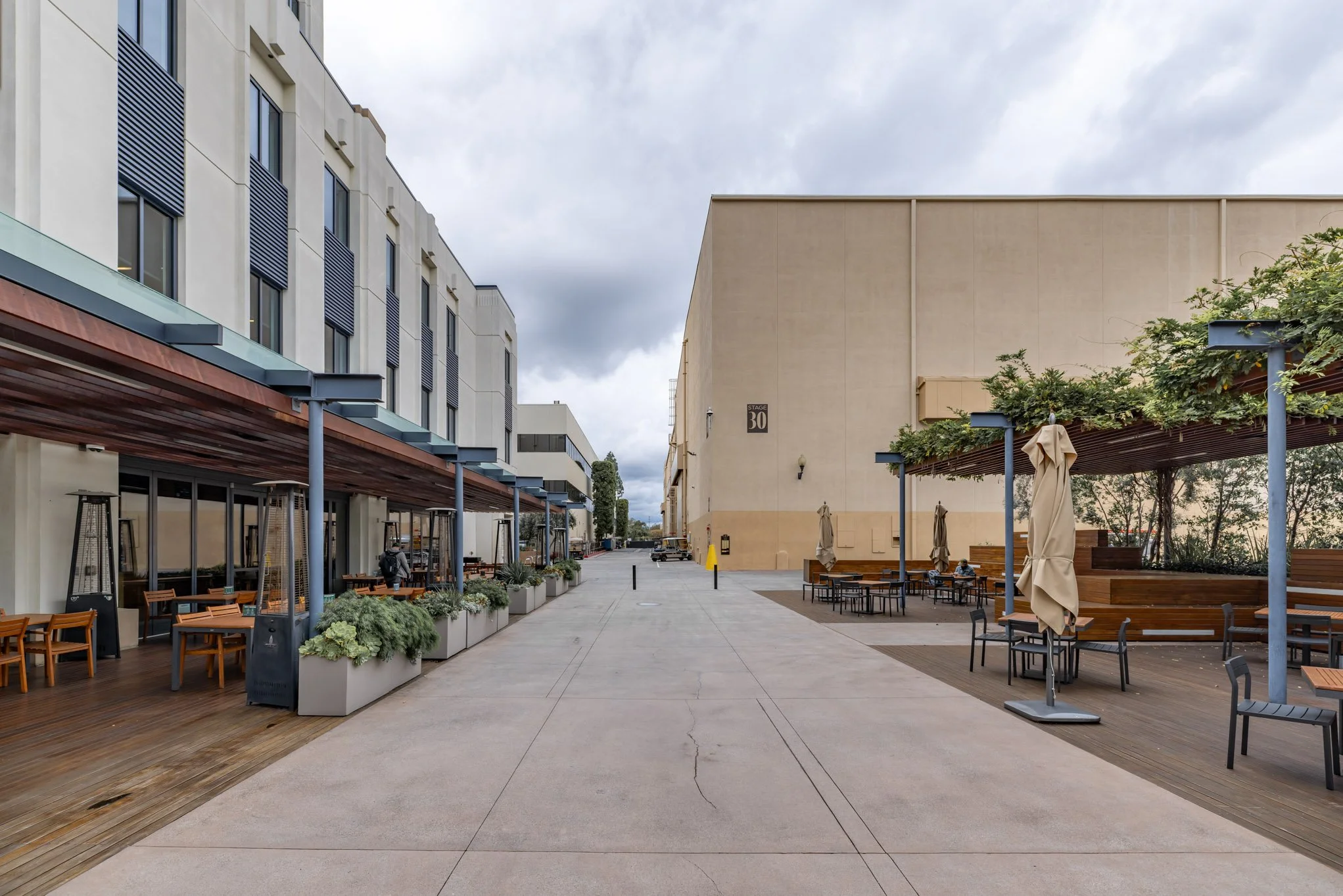 Empty outdoor shopping or dining area with tables, chairs, and umbrellas, bordered by modern buildings, under cloudy sky.