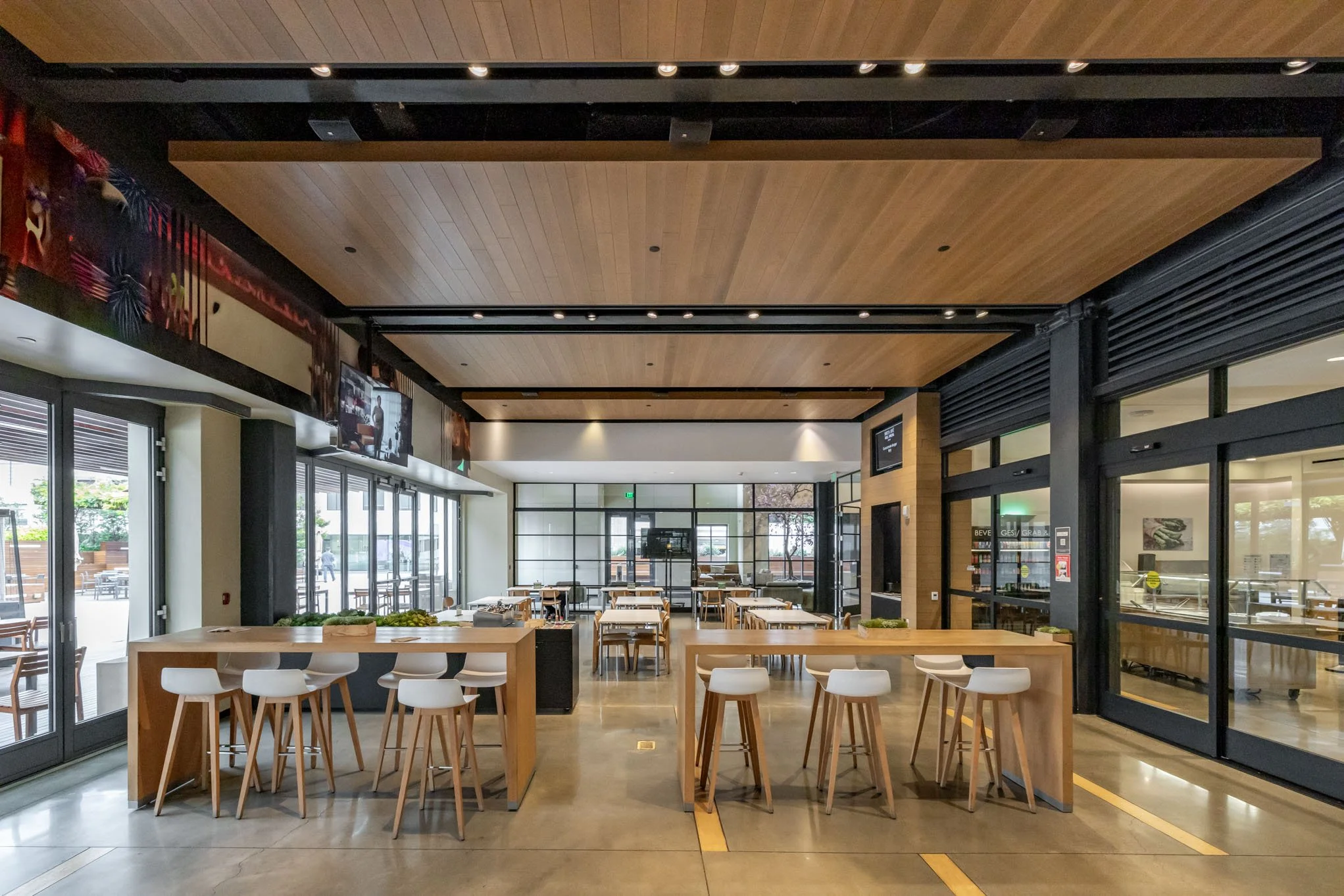 Empty restaurant interior with wooden tables and white chairs, glass doors, and large windows, illuminated by ceiling lights.