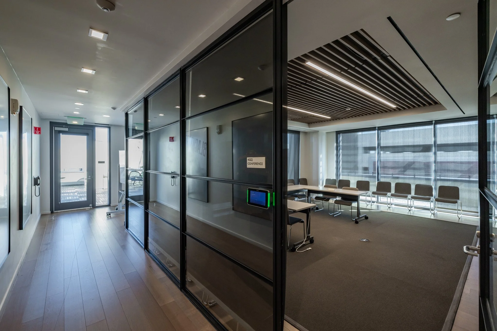 Modern conference room with glass walls, gray chairs arranged around a table, large window with shades, and a ceiling with wooden slats.