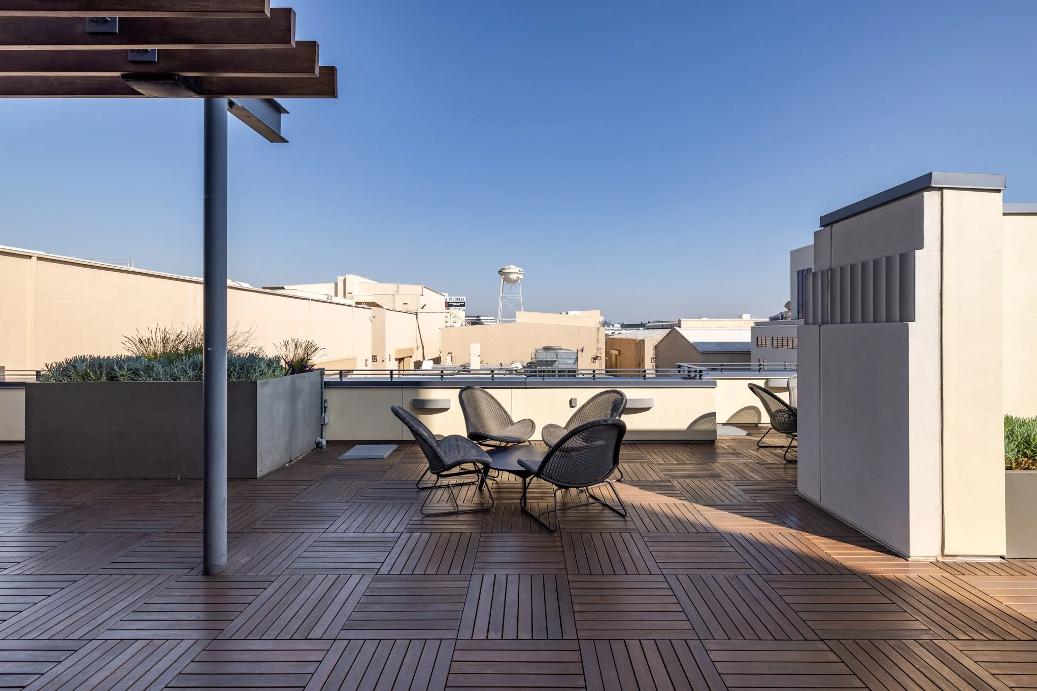 Rooftop terrace with wooden tiles, a round table and four black chairs, planters, and a cityscape with buildings and a water tower under a clear blue sky.