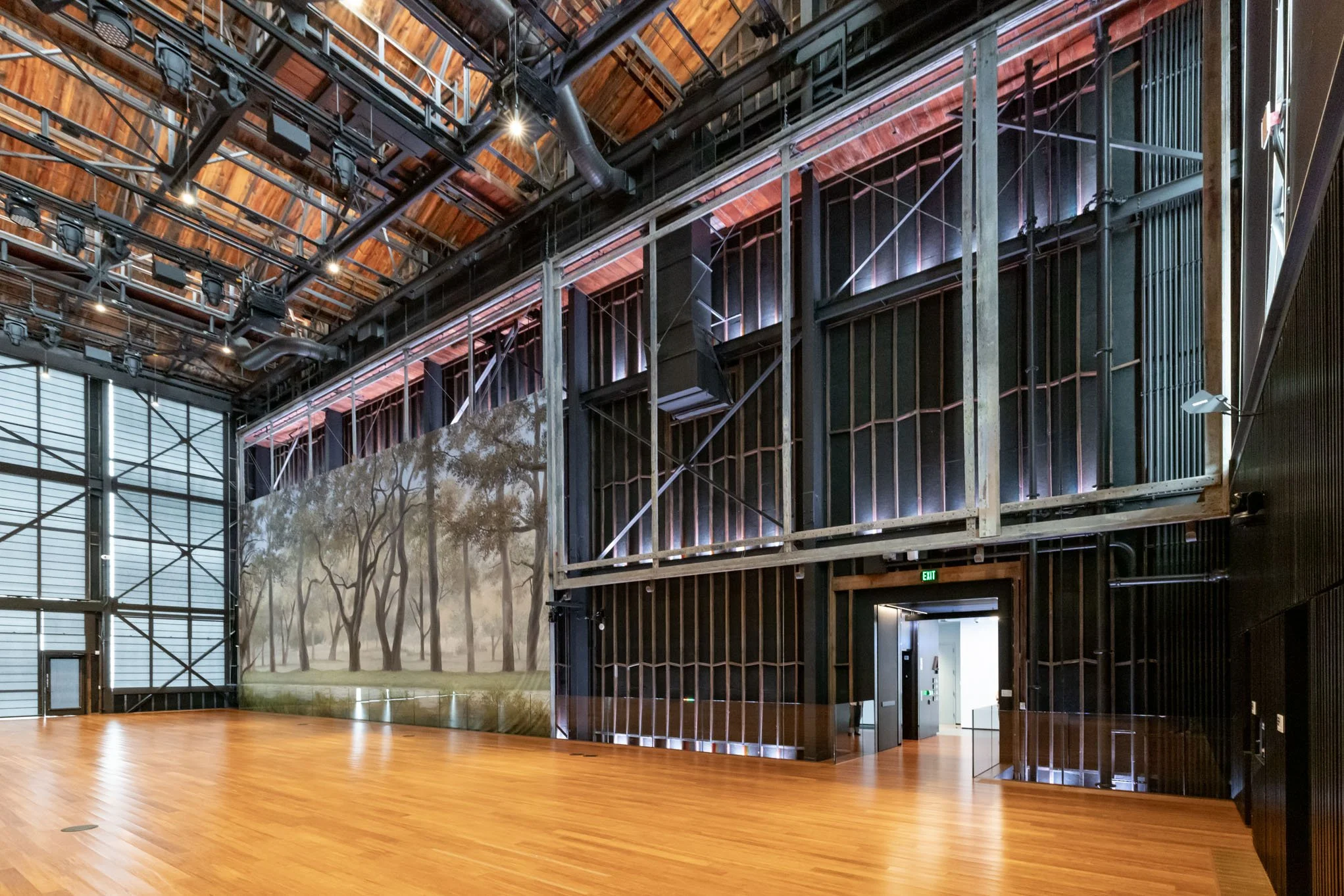 Empty interior space with wooden floor, wall mural of trees and grass, and industrial-style black metal framework with lighting fixtures on the ceiling.