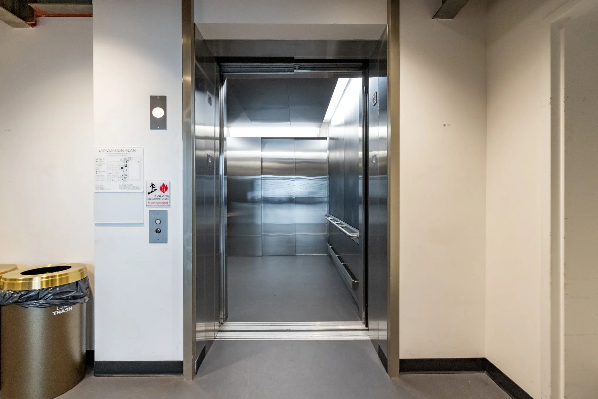 Stainless steel elevator with open doors in a building lobby. There is a trash can to the left and an evacuation plan on the wall.